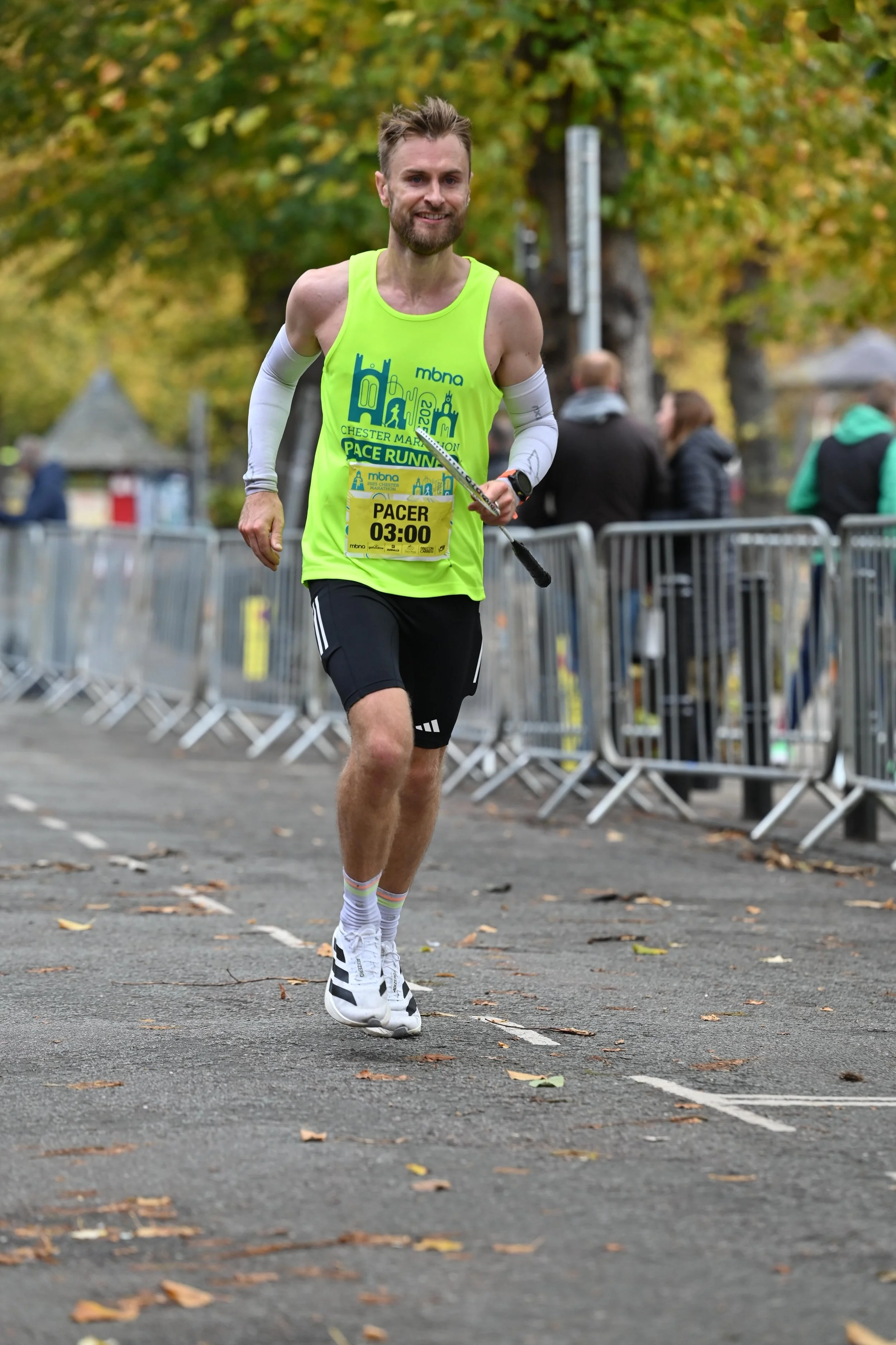 A male marathon runner wearing a neon yellow race bib labeled 'PACER 03:00' and a bright green sleeveless shirt, running on a paved street with autumn foliage and spectators behind metal barriers.
