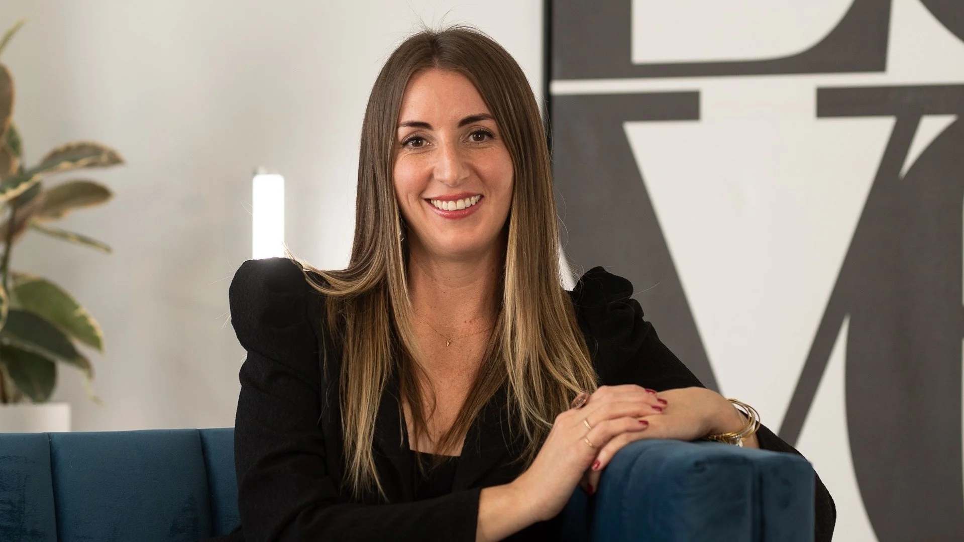 Smiling woman sitting on a blue couch, wearing a black top, with long brown hair, indoors with a modern art background.