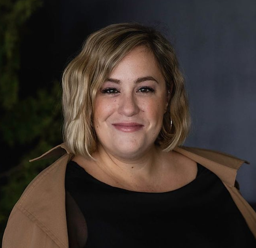 Person with short blond hair smiling, wearing a black top and tan jacket.