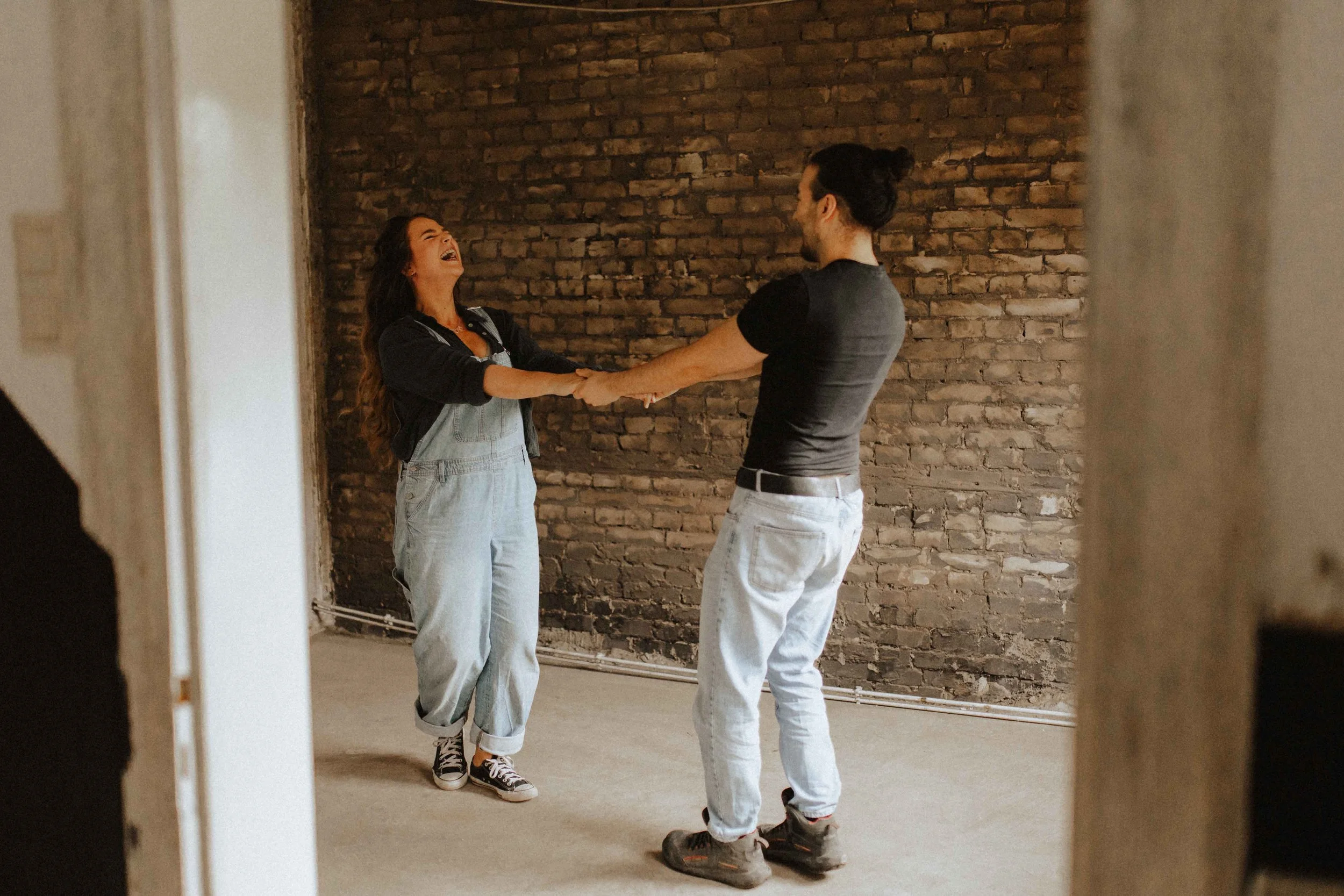 Verspieltes Paarshooting in Lüneburg – lachendes Paar hält sich an den Händen und tanzt vor einer Backsteinwand, eingefangen von einer kreativen Paarfotografin.
