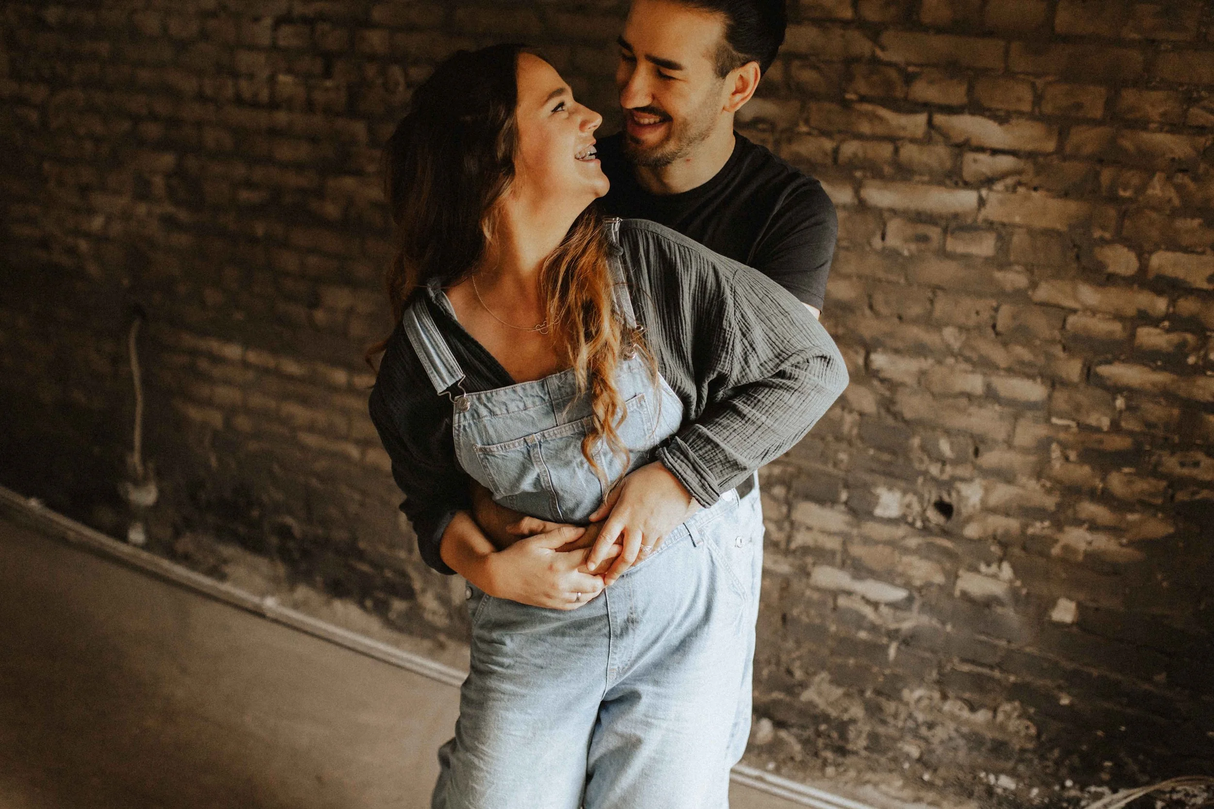 Verspieltes Paarshooting in Lüneburg – Mann umarmt seine Partnerin von hinten, beide lachen herzlich vor einer rustikalen Backsteinwand.