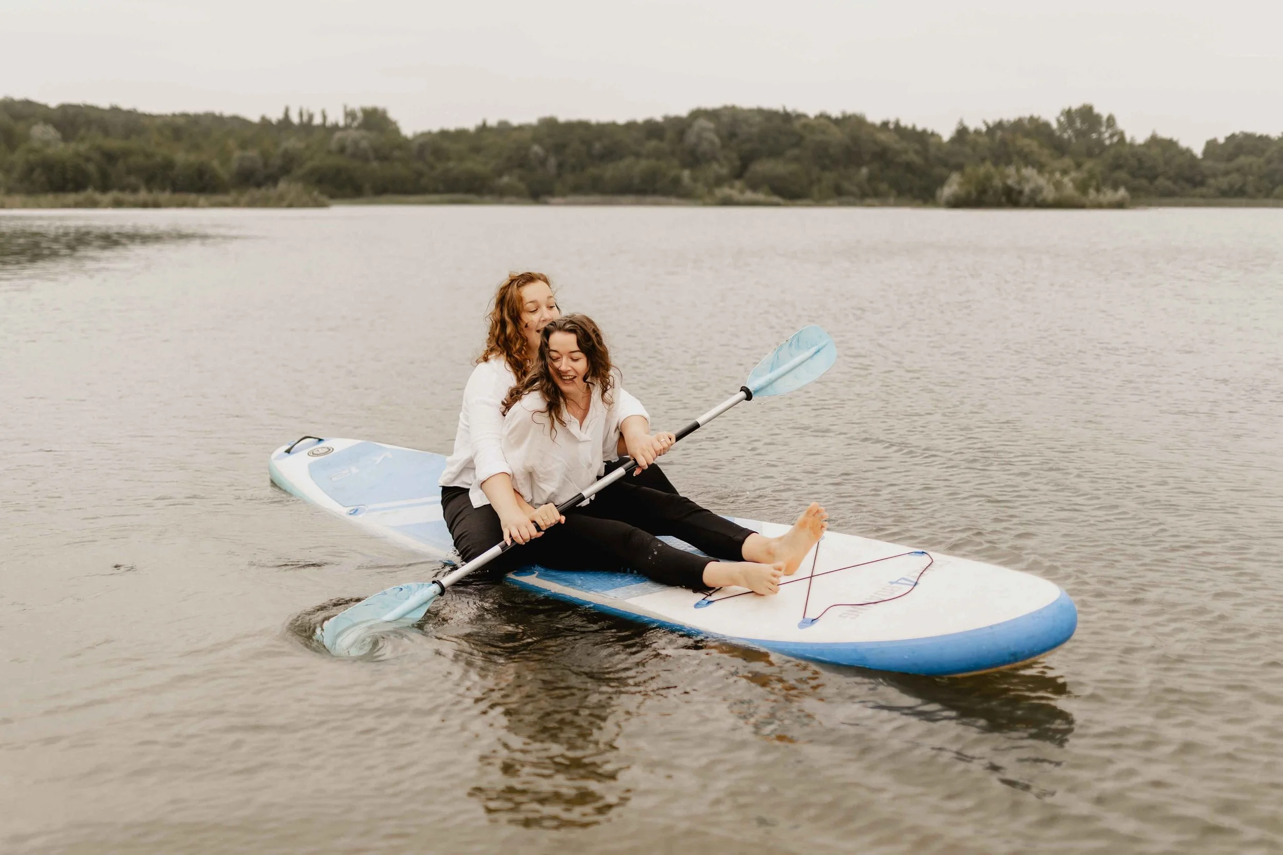 Zwei Frauen sitzen gemeinsam auf einem SUP-Board auf dem See und halten lachend ein Paddel