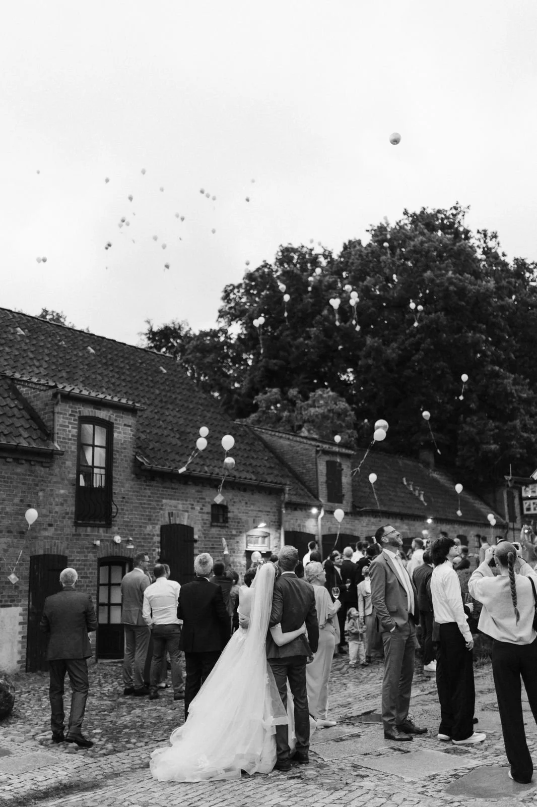 Schwarz-Weiß-Aufnahme einer Hochzeitsgesellschaft im Innenhof des Heide Hotels Reinstorf bei Lüneburg. Das Brautpaar steht eng beieinander, während die Gäste den aufsteigenden Luftballons über den historischen Backsteingebäuden nachschauen.