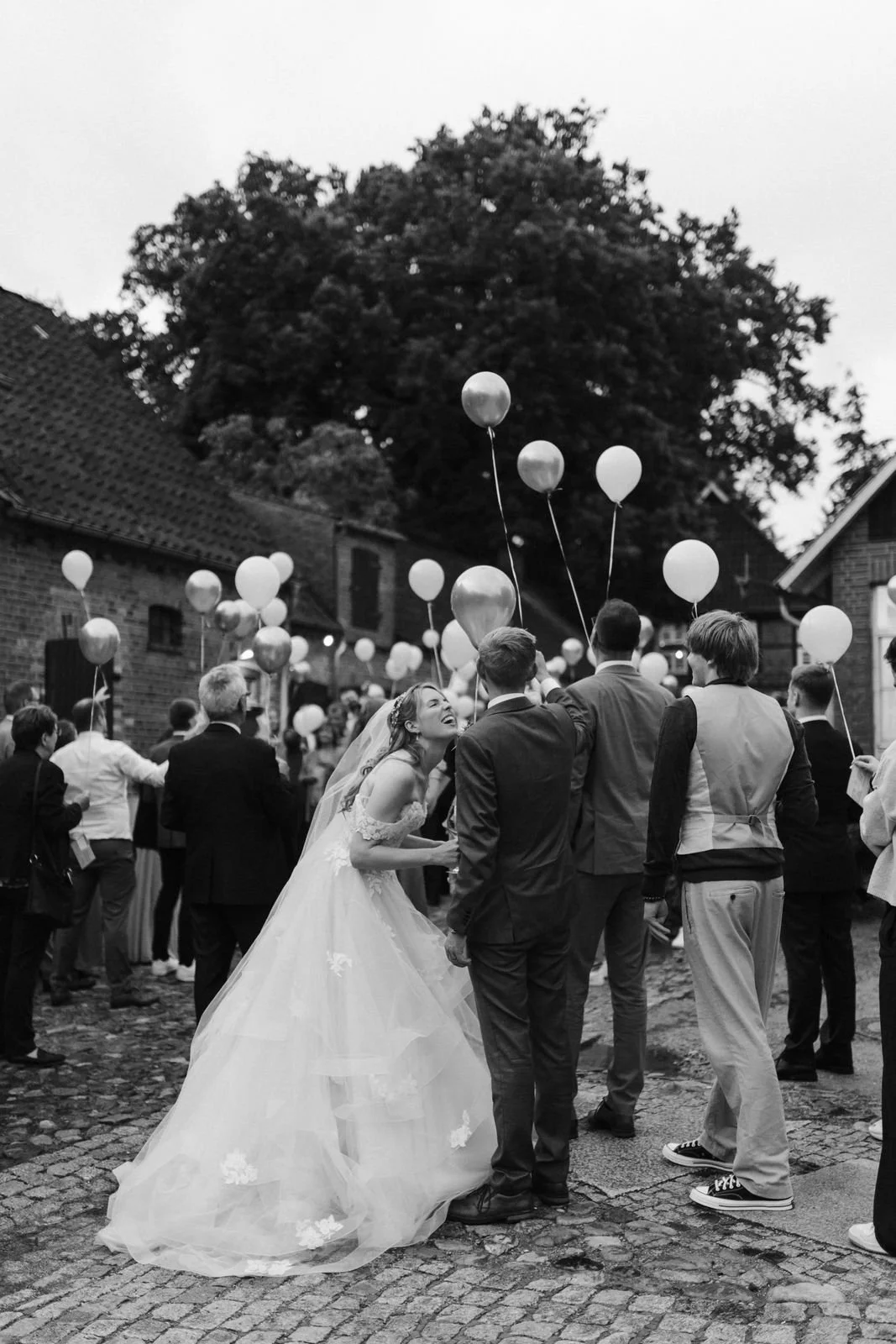 Schwarz-Weiß-Aufnahme einer lachenden Braut inmitten ihrer Hochzeitsgäste mit zahlreichen Luftballons im Innenhof des Heide Hotels Reinstorf bei Lüneburg.