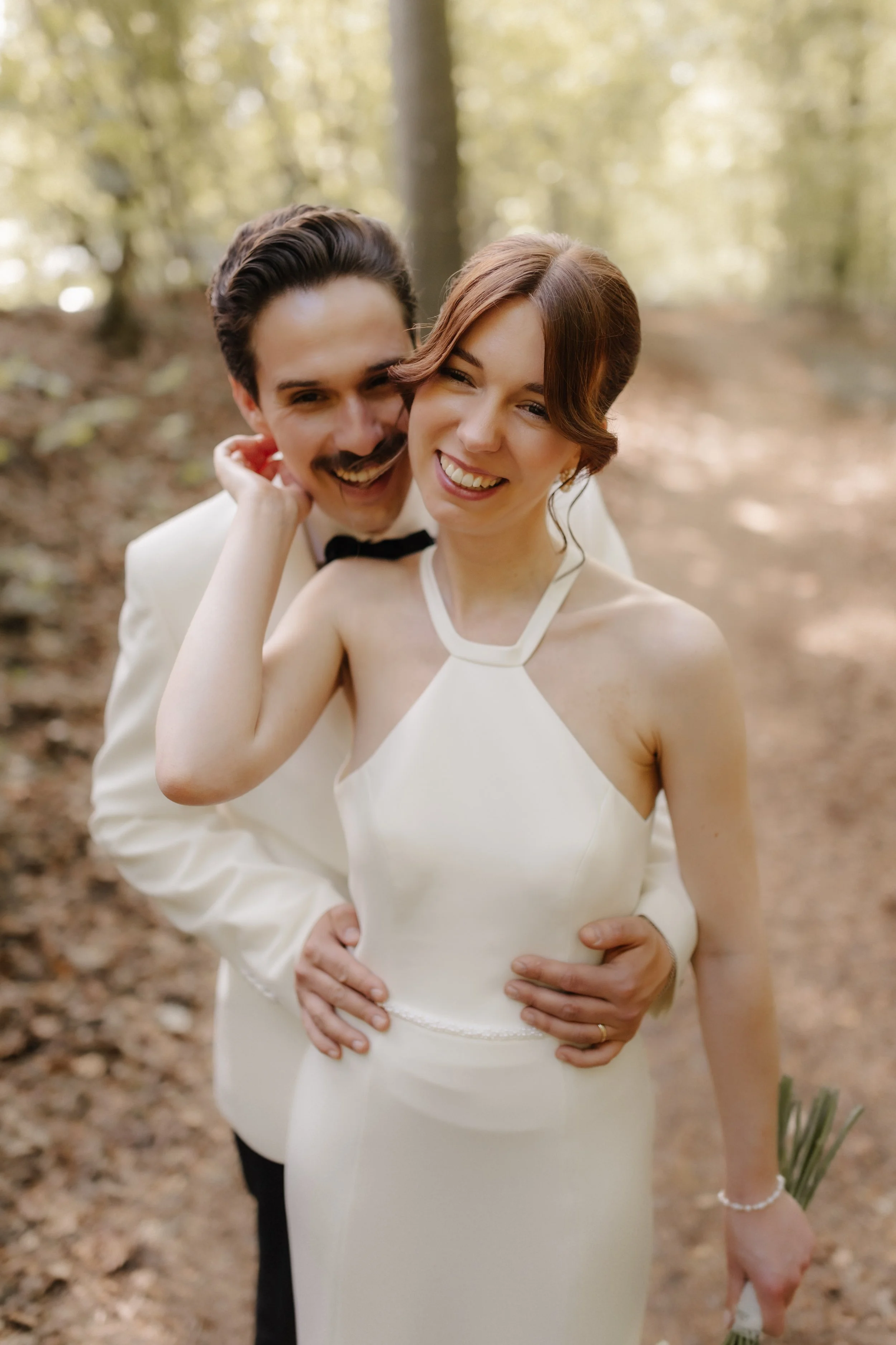 Glückliches Brautpaar bei einem romantischen Hochzeitsshooting im Wald bei Lüneburg. Der Bräutigam umarmt seine lachende Braut liebevoll von hinten.