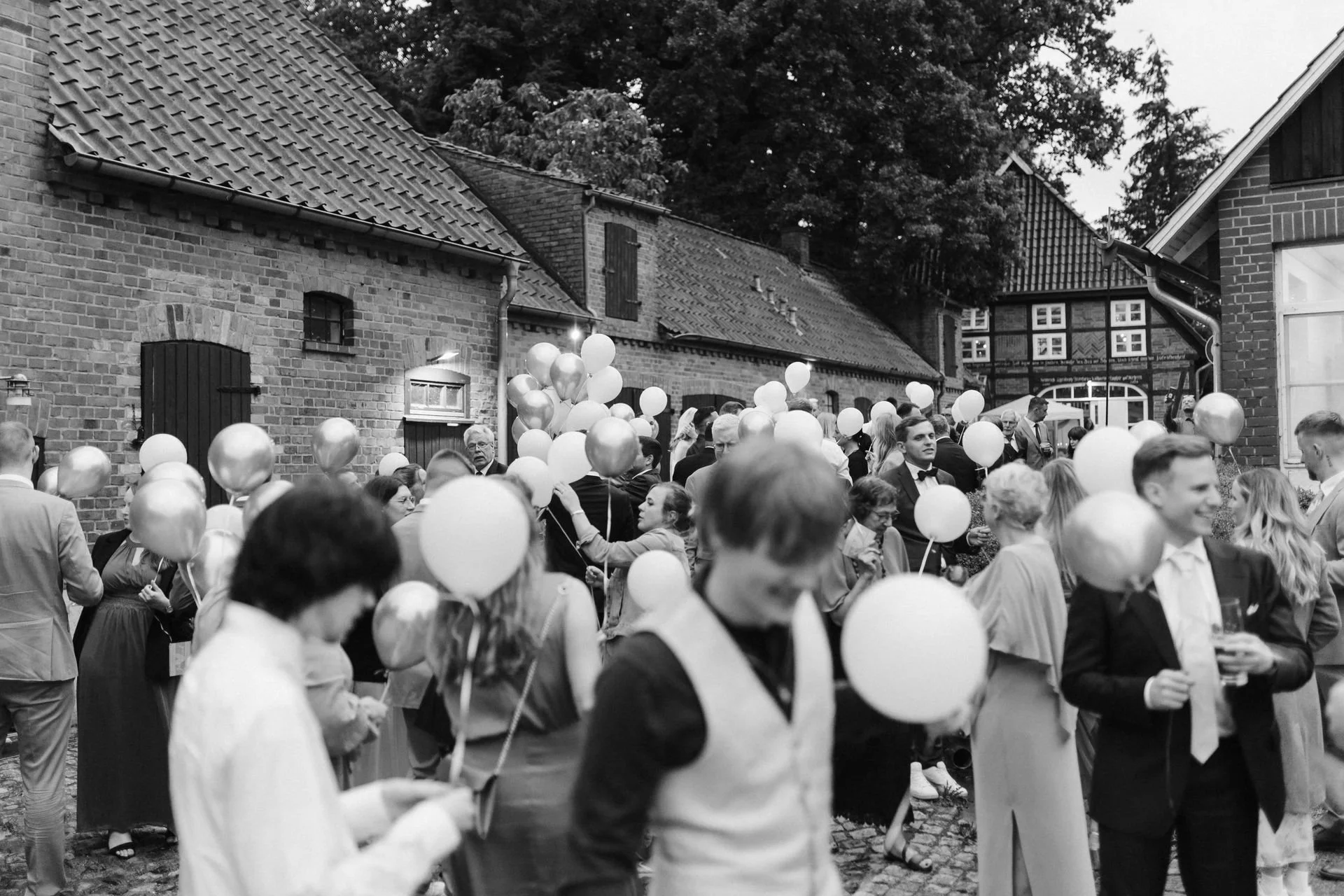 Schwarz-Weiß-Aufnahme einer Hochzeitsgesellschaft im Innenhof des Heide Hotels Reinstorf bei Lüneburg. Zahlreiche Hochzeitsgäste versammeln sich zwischen den historischen Backsteingebäuden und halten Luftballons in den Händen.