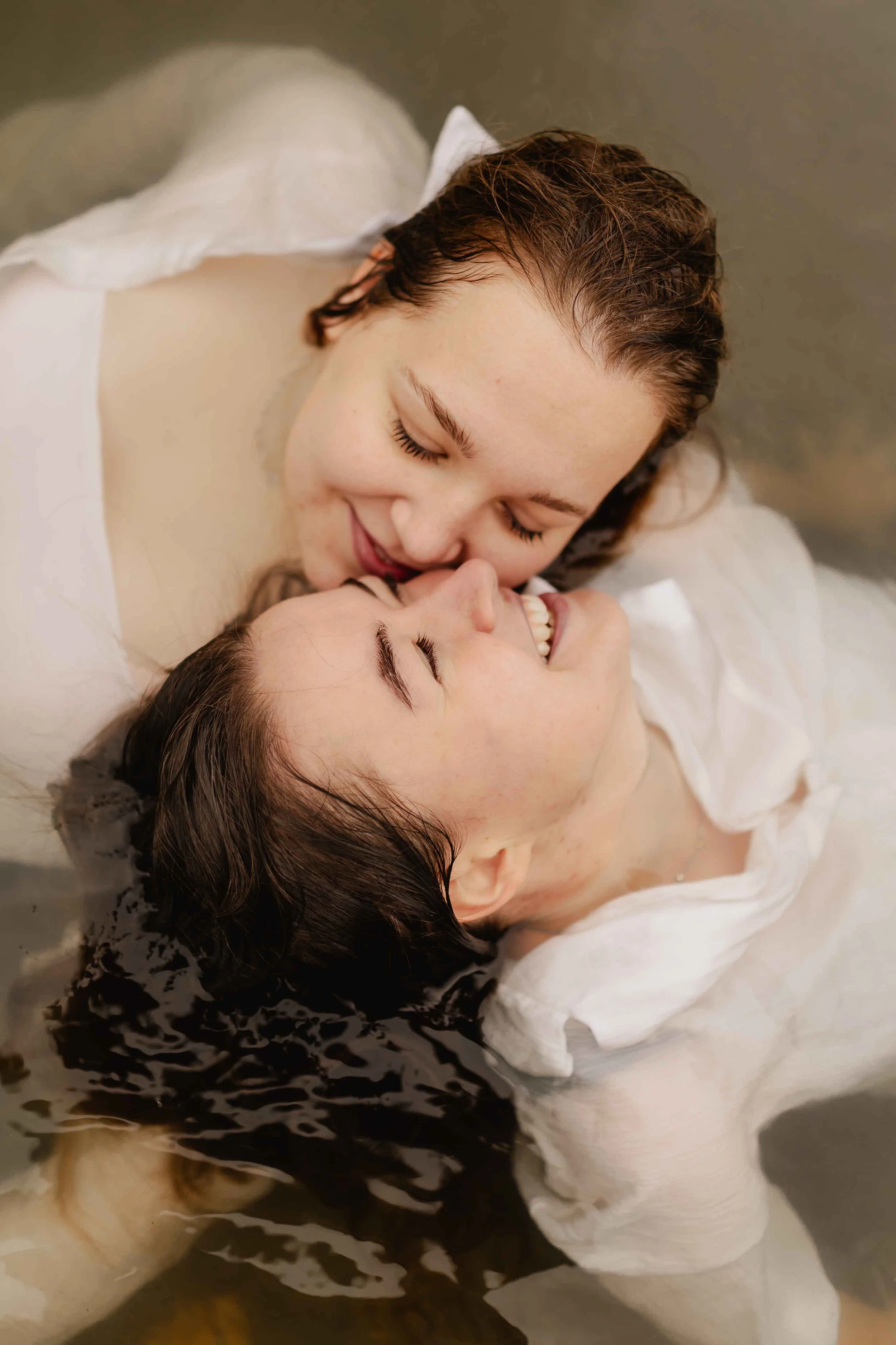 Zwei Frauen liegen eng umschlungen im Wasser, eine Partnerin lacht nach oben, während die andere sie liebevoll hält