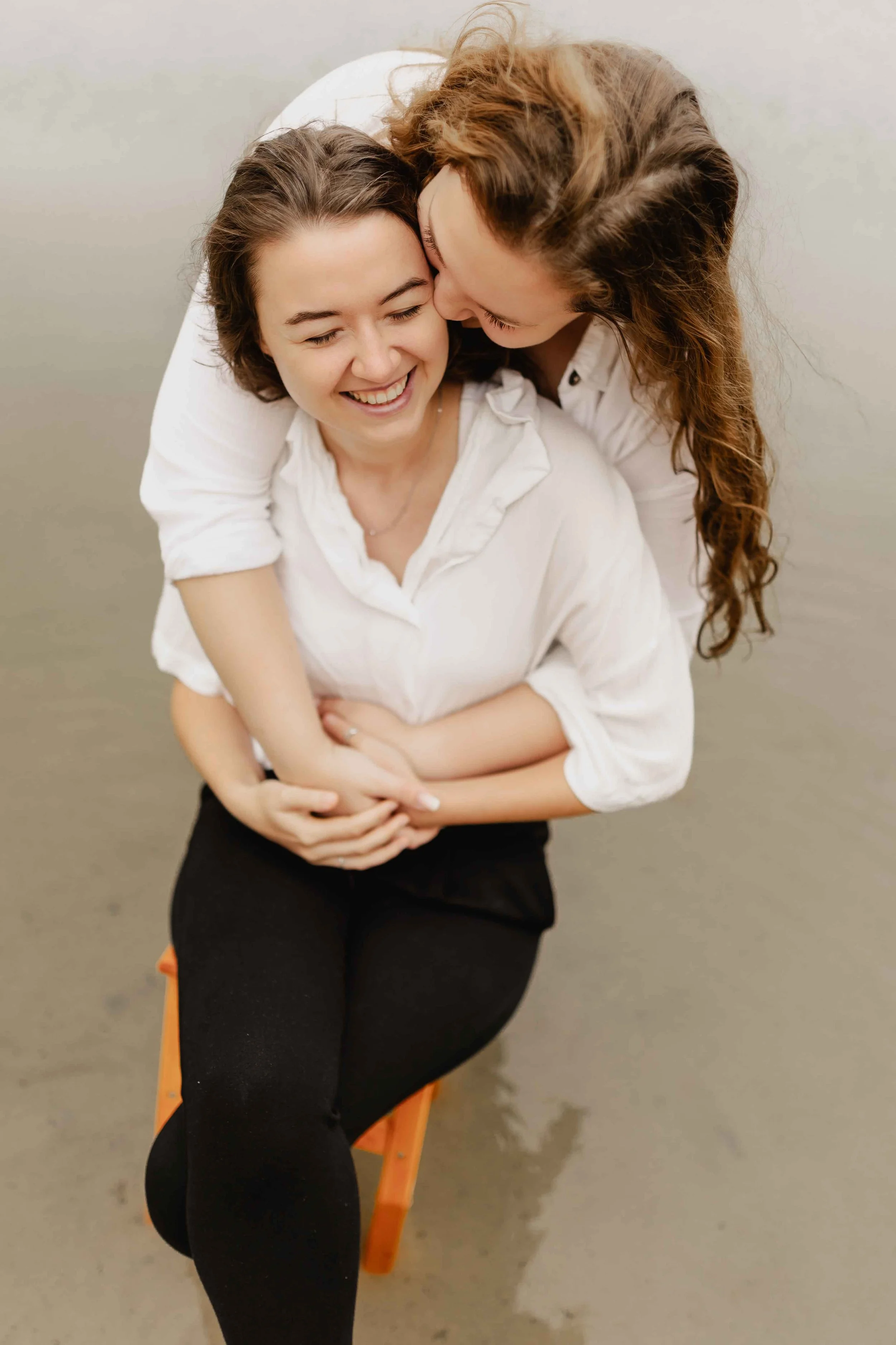 Frau sitzt lachend auf einem Hocker im seichten Wasser, ihre Partnerin umarmt sie von hinten und küsst sie auf die Wange