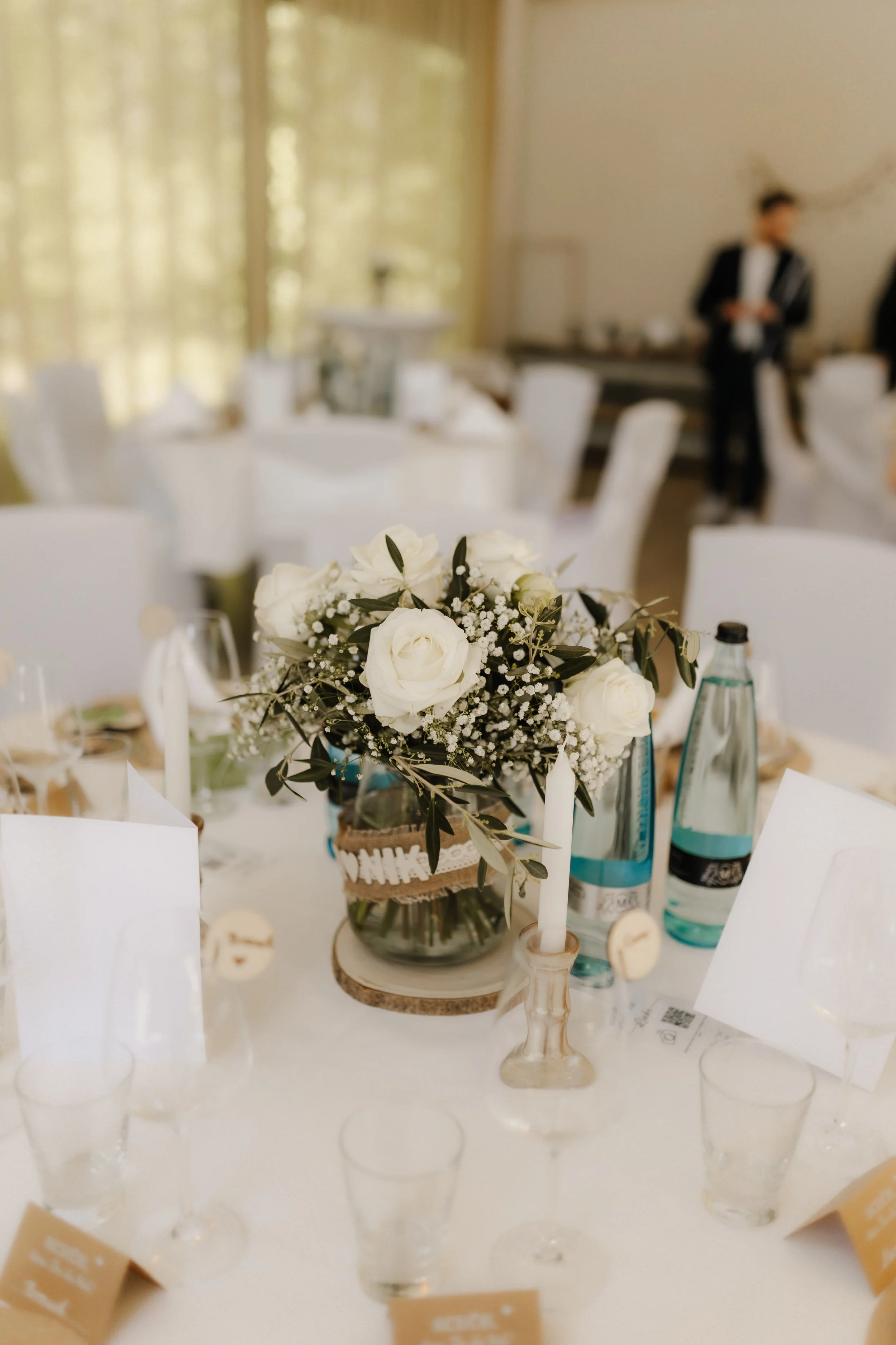 Elegante Tischdekoration bei einer Hochzeit mit weißen Rosen, Schleierkraut und Olivenzweigen in einer Glasvase auf einer festlich gedeckten Tafel.