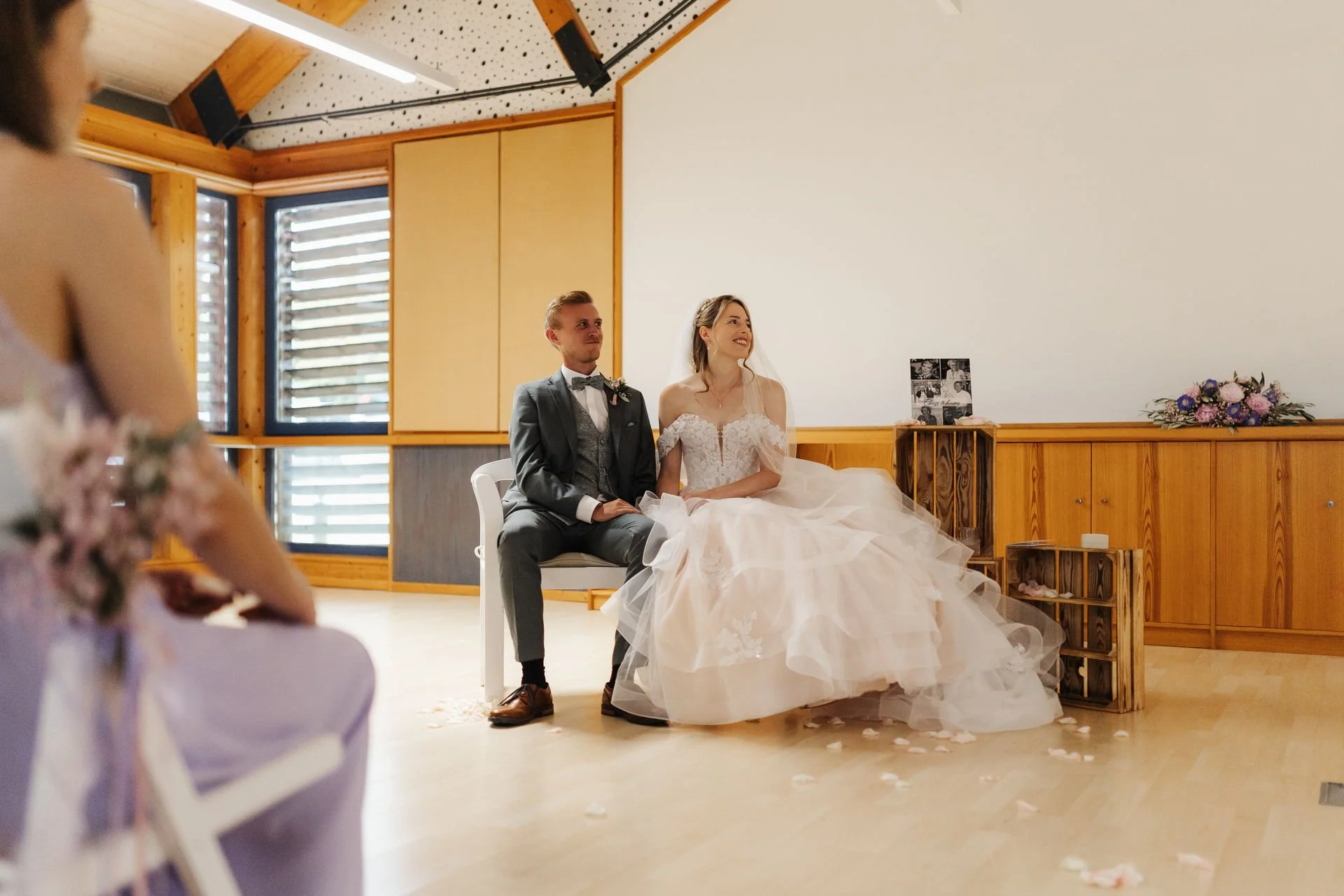 Brautpaar Corinna und Lukas sitzt lächelnd während seiner freien Trauung im Heide Hotel Reinstorf bei Lüneburg. Die Braut trägt ein romantisches Hochzeitskleid mit langer Schleppe, der Bräutigam einen grauen Anzug.