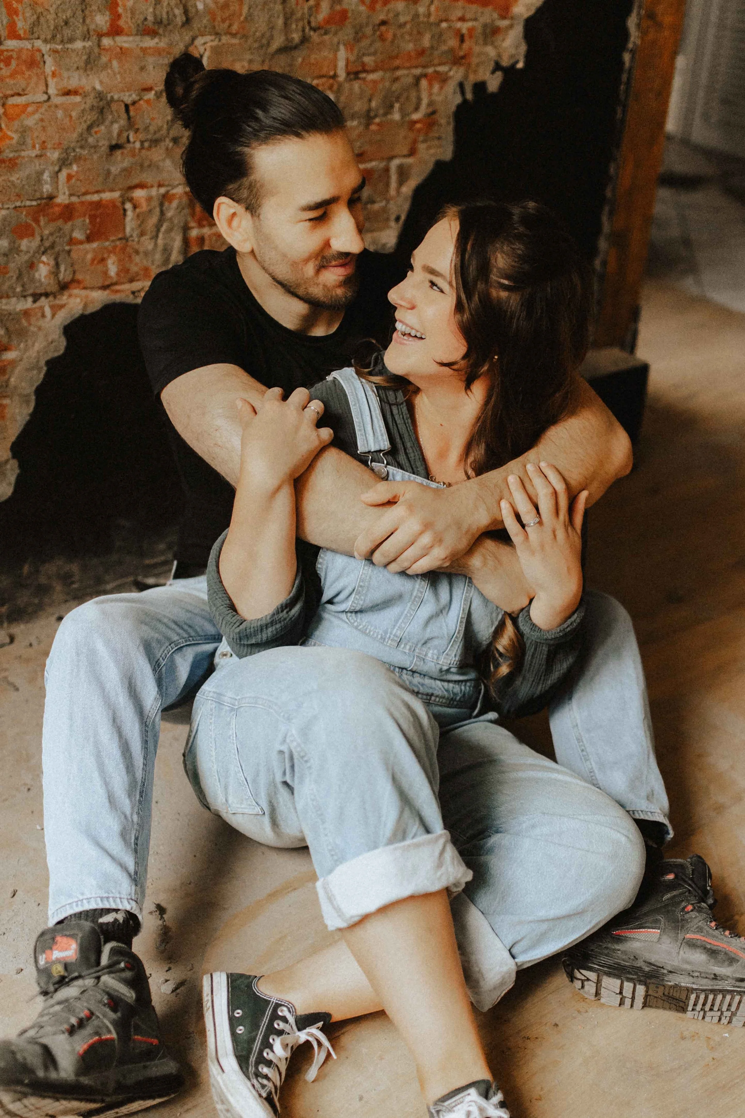Paarshooting in Lüneburg – verliebtes Paar sitzt auf dem Boden vor einer Backsteinwand, hält sich innig im Arm und lacht sich an.