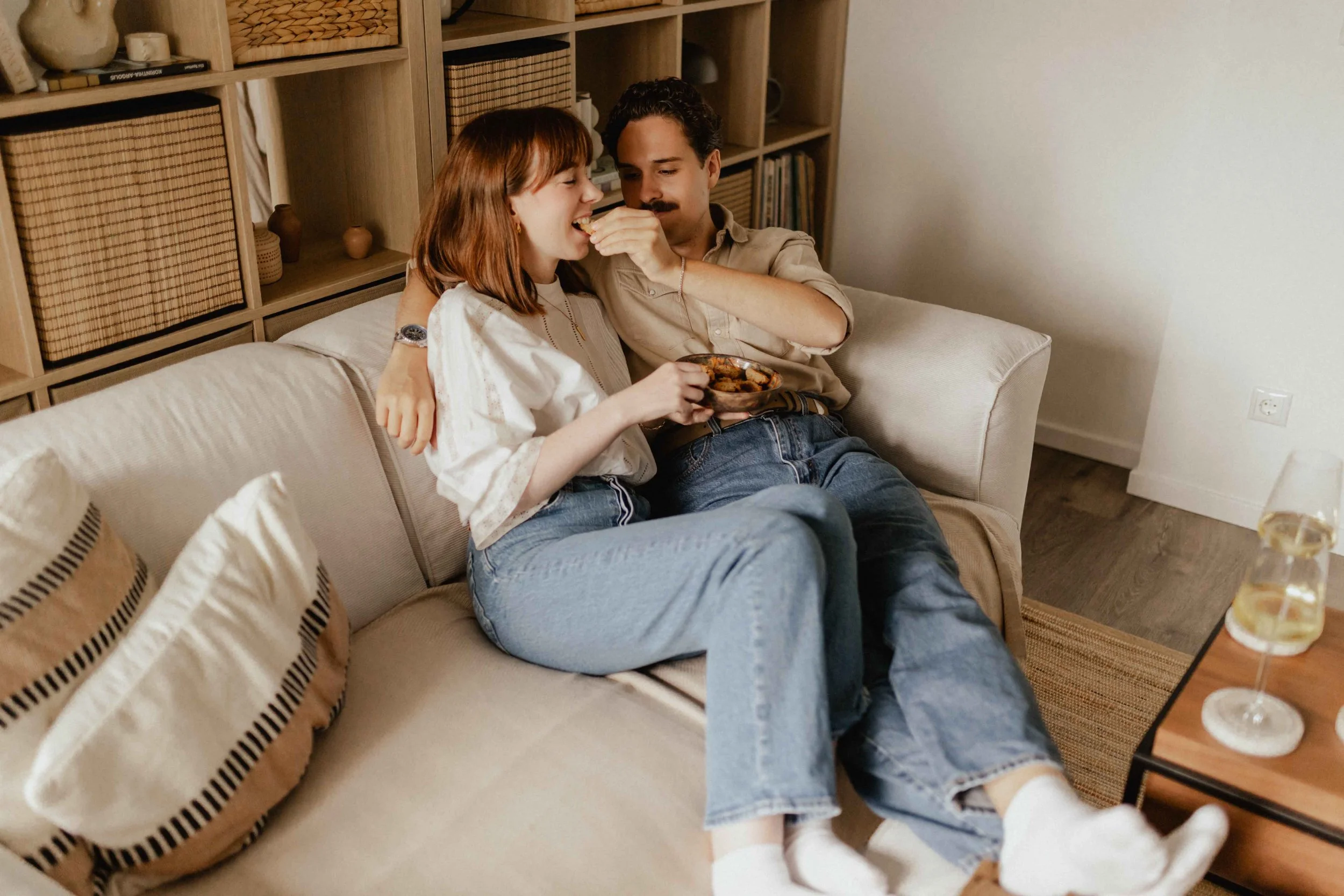 Paar sitzt bei einer Homestory in Lüneburg eng umschlungen auf dem Sofa, isst Gebäck und trinkt Wein in entspannter Atmosphäre