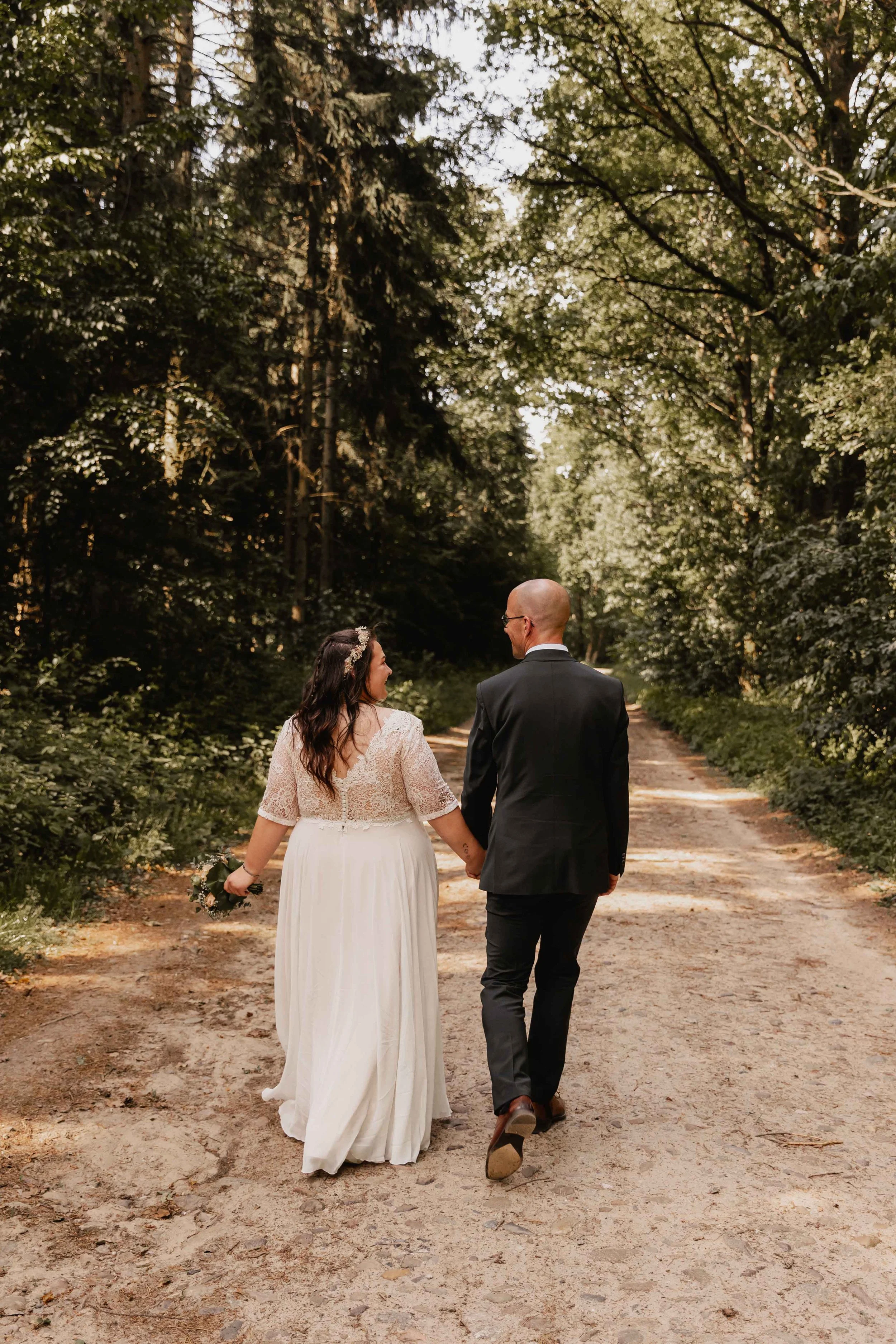 Brautpaar spaziert Hand in Hand einen Waldweg entlang in entspannter Stimmung