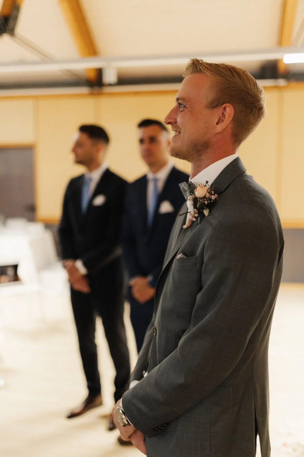 Lächelnder Bräutigam im grauen Hochzeitsanzug mit Blumenanstecker wartet gemeinsam mit seinen Trauzeugen auf die Braut bei der Hochzeitsfeier im Heide Hotel Reinstorf bei Lüneburg.