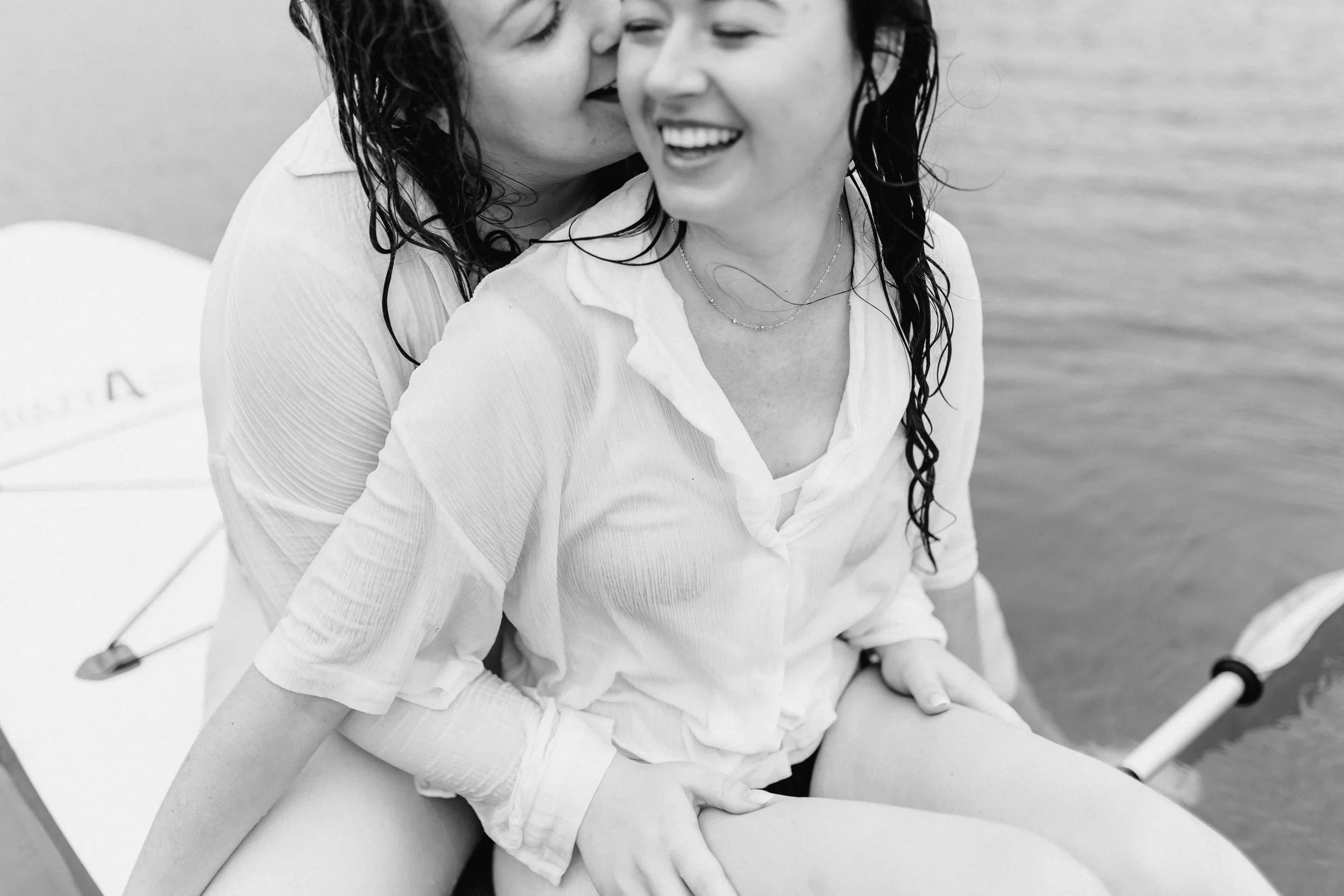 Zwei Frauen sitzen auf einem SUP-Board, die hintere Partnerin umarmt ihre Liebste von hinten und beide lachen glücklich