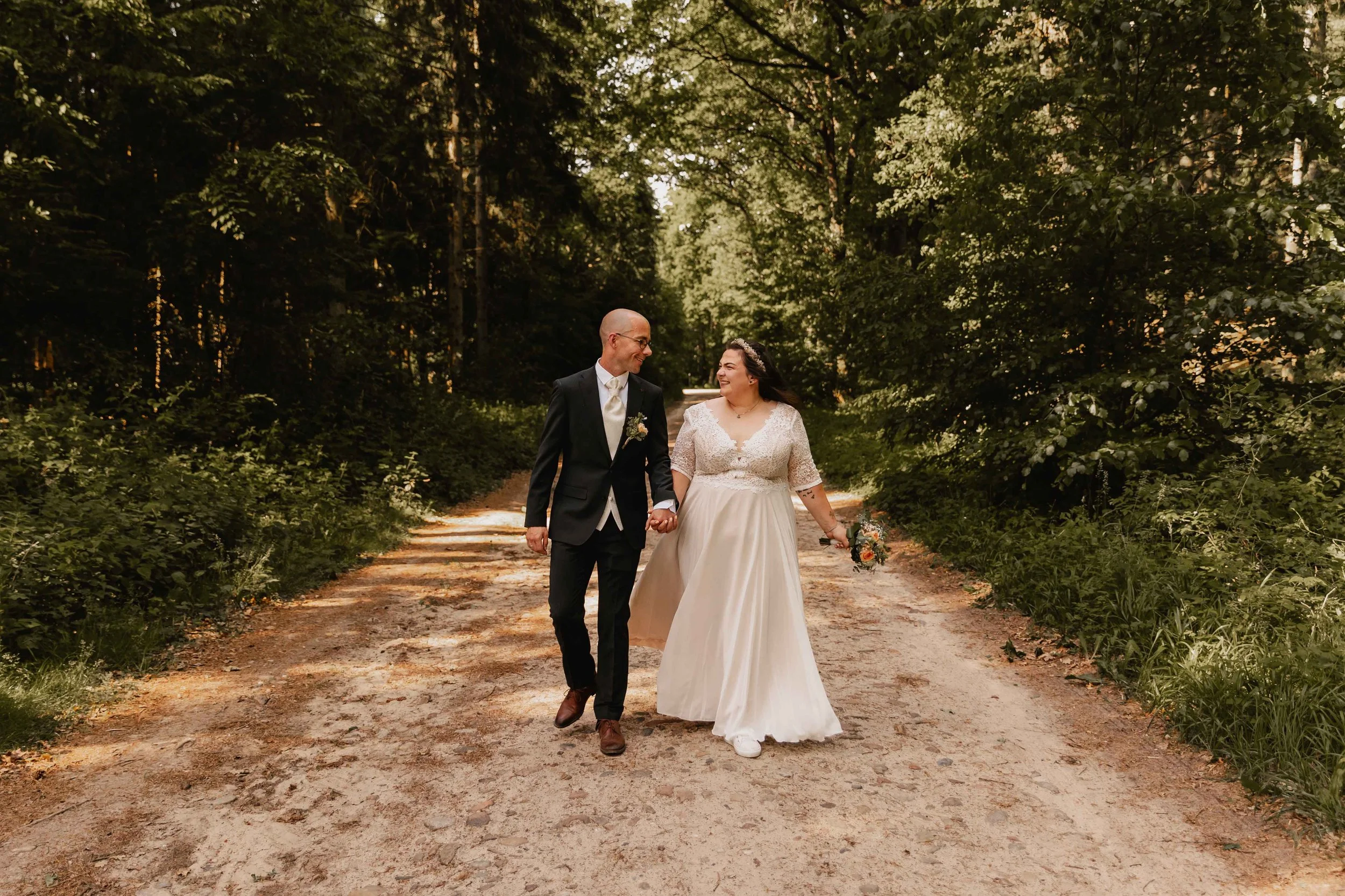 Hochzeitspaar läuft lachend nebeneinander über einen Waldweg bei Sonnenschein