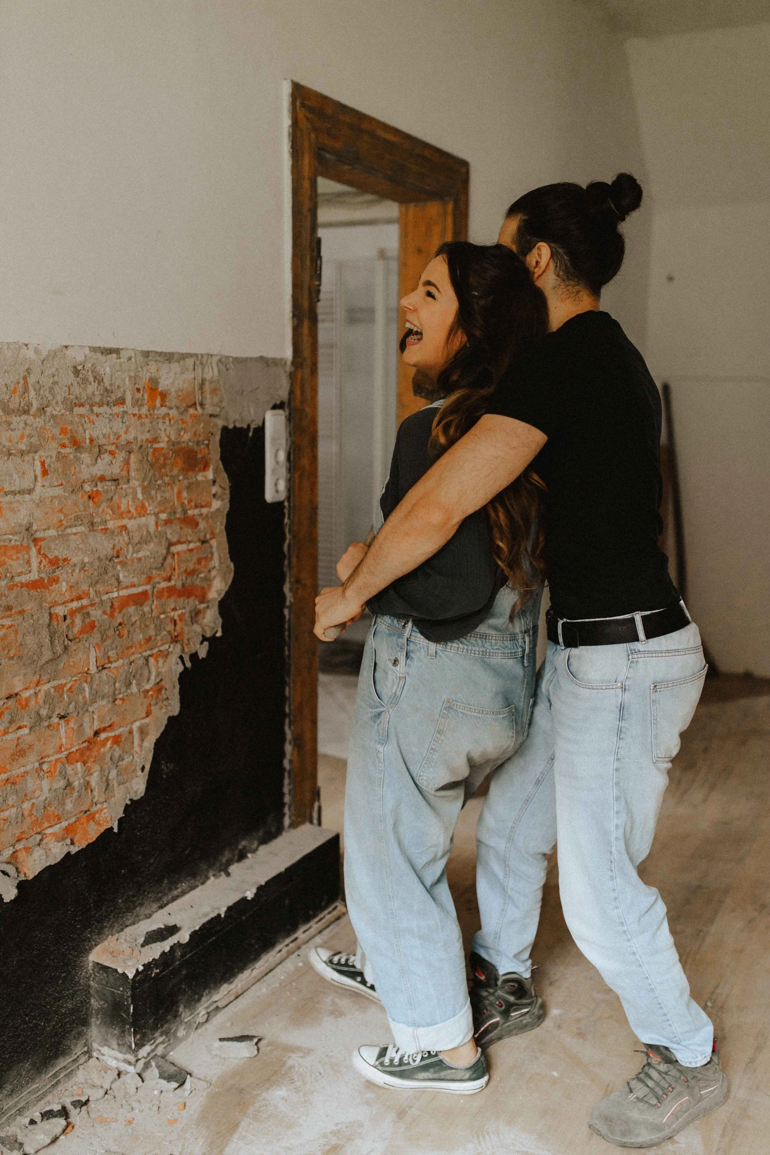 Authentisches Paarshooting in Lüneburg – Mann umarmt seine lachende Partnerin von hinten in einer industriellen Loft-Location mit Backsteinwand.