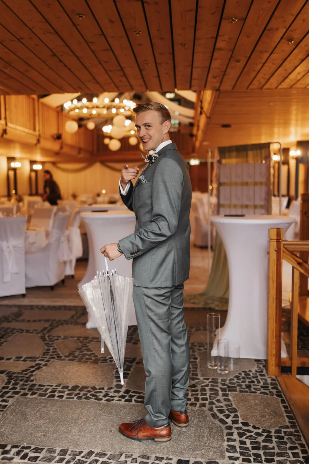 Lächelnder Bräutigam im eleganten grauen Hochzeitsanzug mit Fliege und transparentem Regenschirm im festlich dekorierten Hochzeitssaal des Heide Hotels Reinstorf bei Lüneburg.