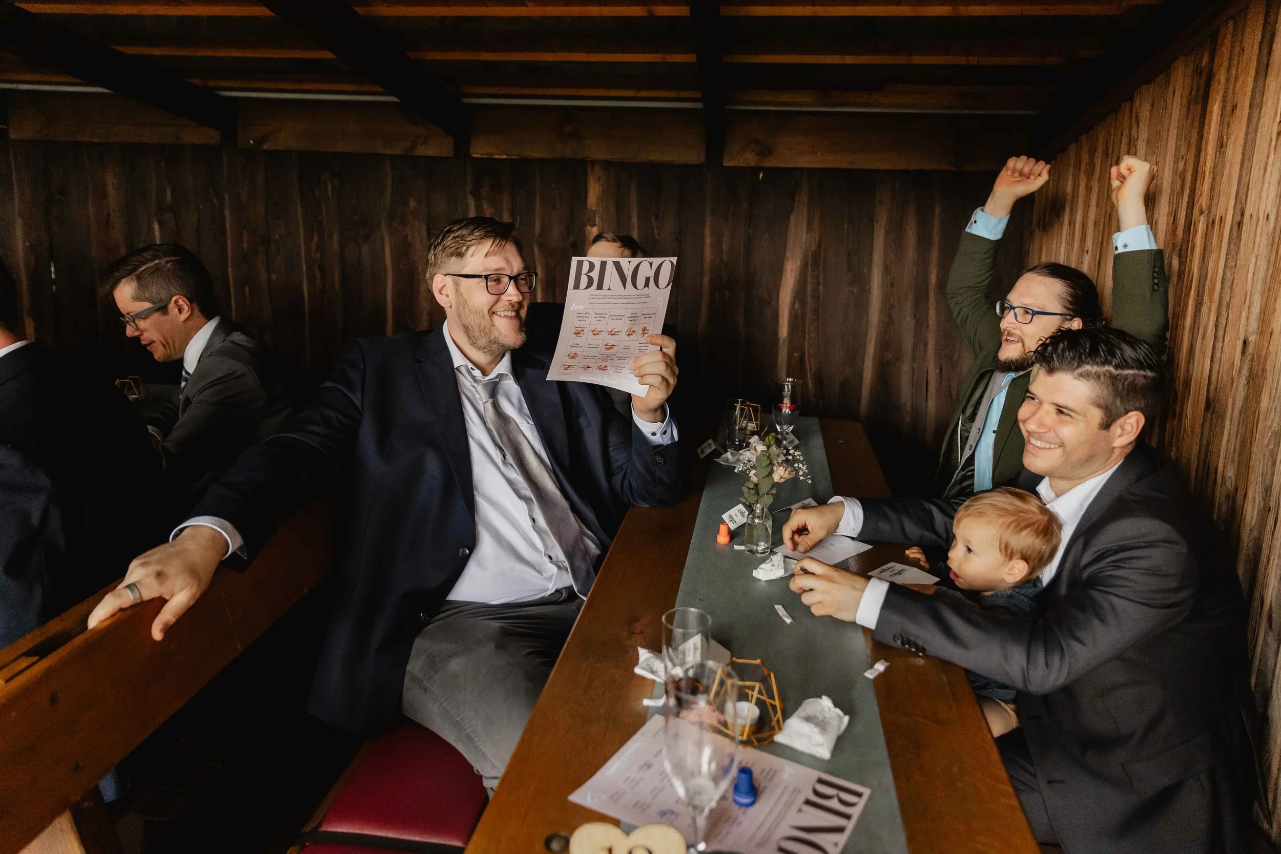 Gäste jubeln bei einer Bingo-Spielrunde mit Getränken und Kind auf dem Schoß