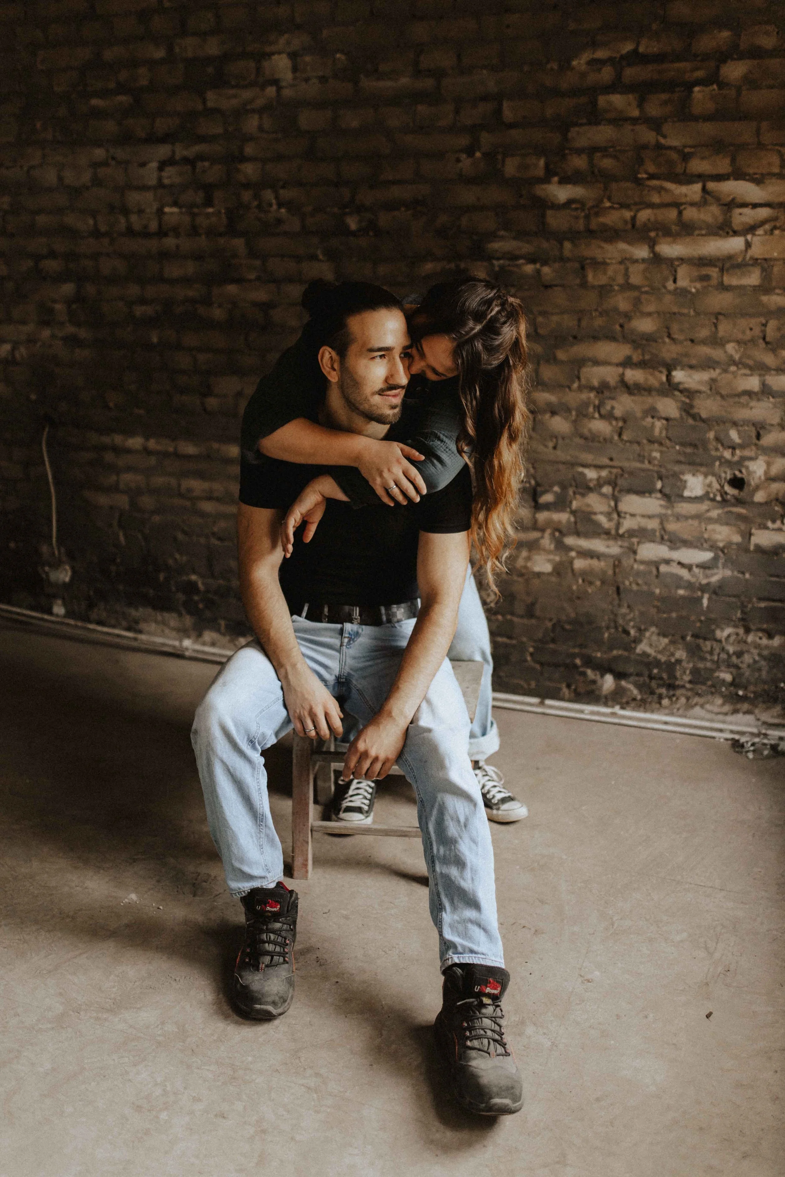 Paarshooting in Lüneburg – Frau umarmt ihren sitzenden Partner liebevoll von hinten vor einer rustikalen Backsteinwand.