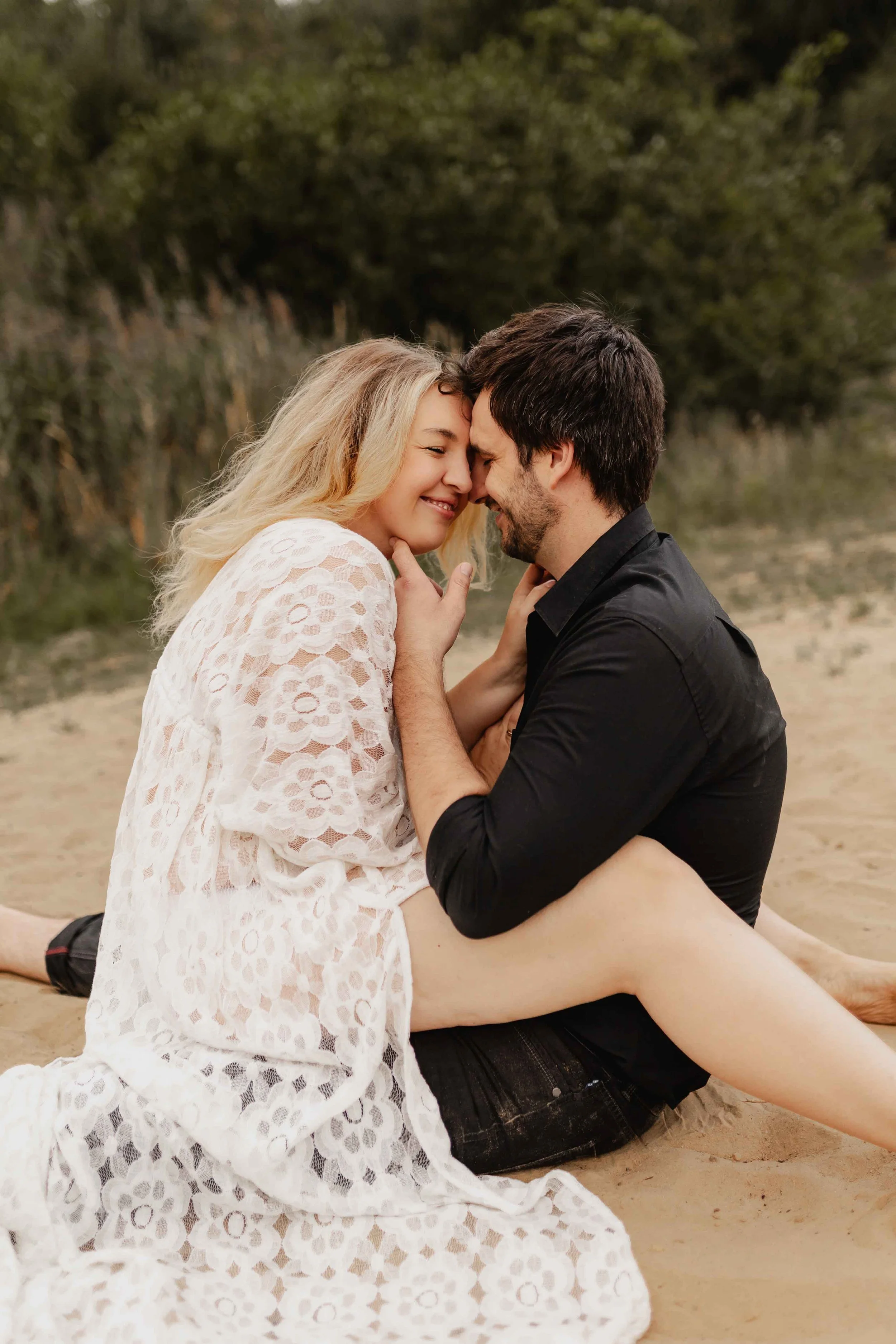 Paar sitzt eng umschlungen am Ufer, sie berühren sanft ihre Stirn aneinander und lächeln vertraut