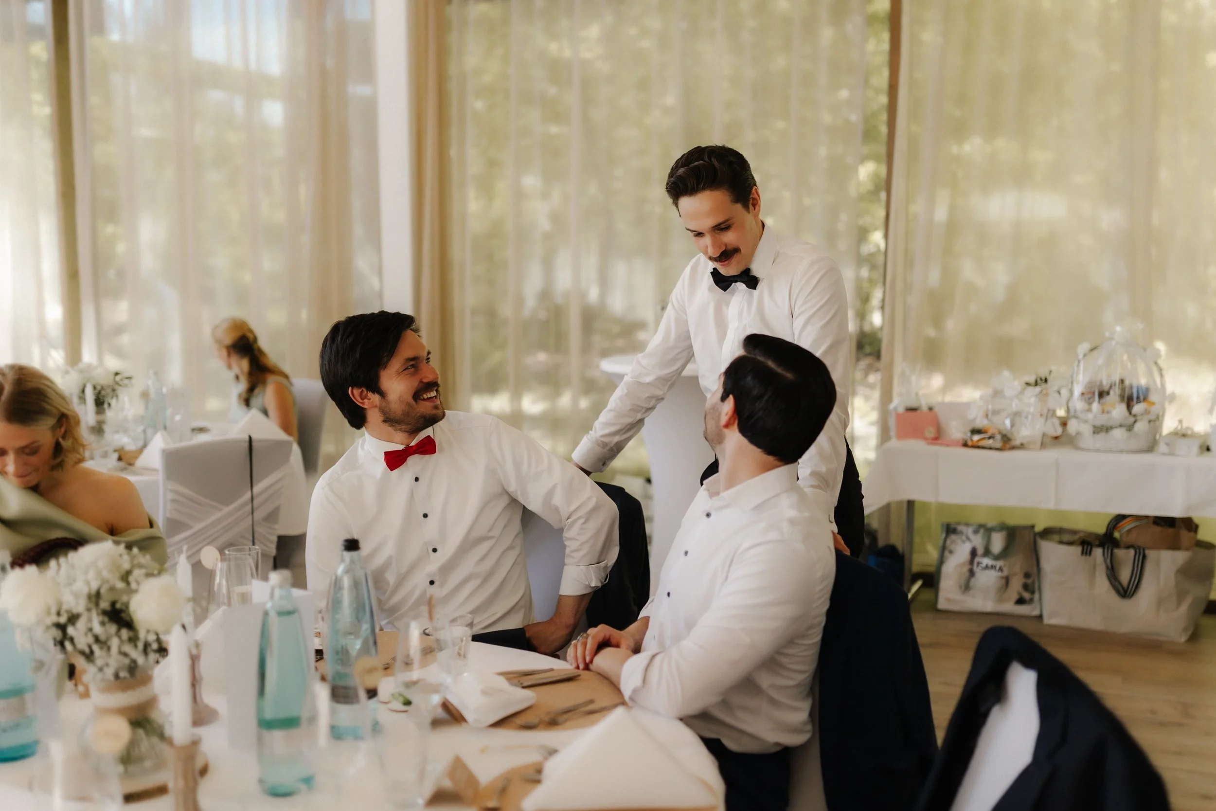 Bräutigam unterhält sich lachend mit zwei Hochzeitsgästen an der festlich gedeckten Tafel im Forsthaus Rote Schleuse in Lüneburg.