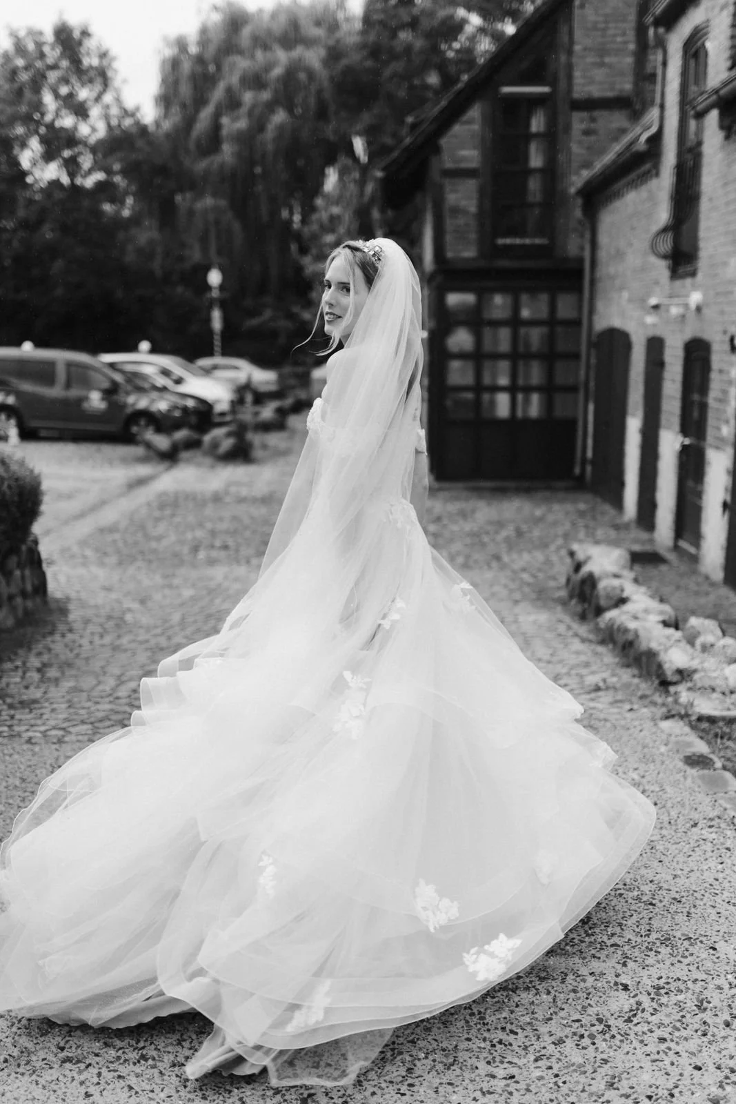 Elegante Schwarz-Weiß-Hochzeitsfotografie einer Braut, die sich in ihrem voluminösen Brautkleid mit langer Schleppe und Schleier umdreht. Romantisches Brautporträt vor dem historischen Backsteingebäude des Heide Hotels Reinstorf bei Lüneburg.