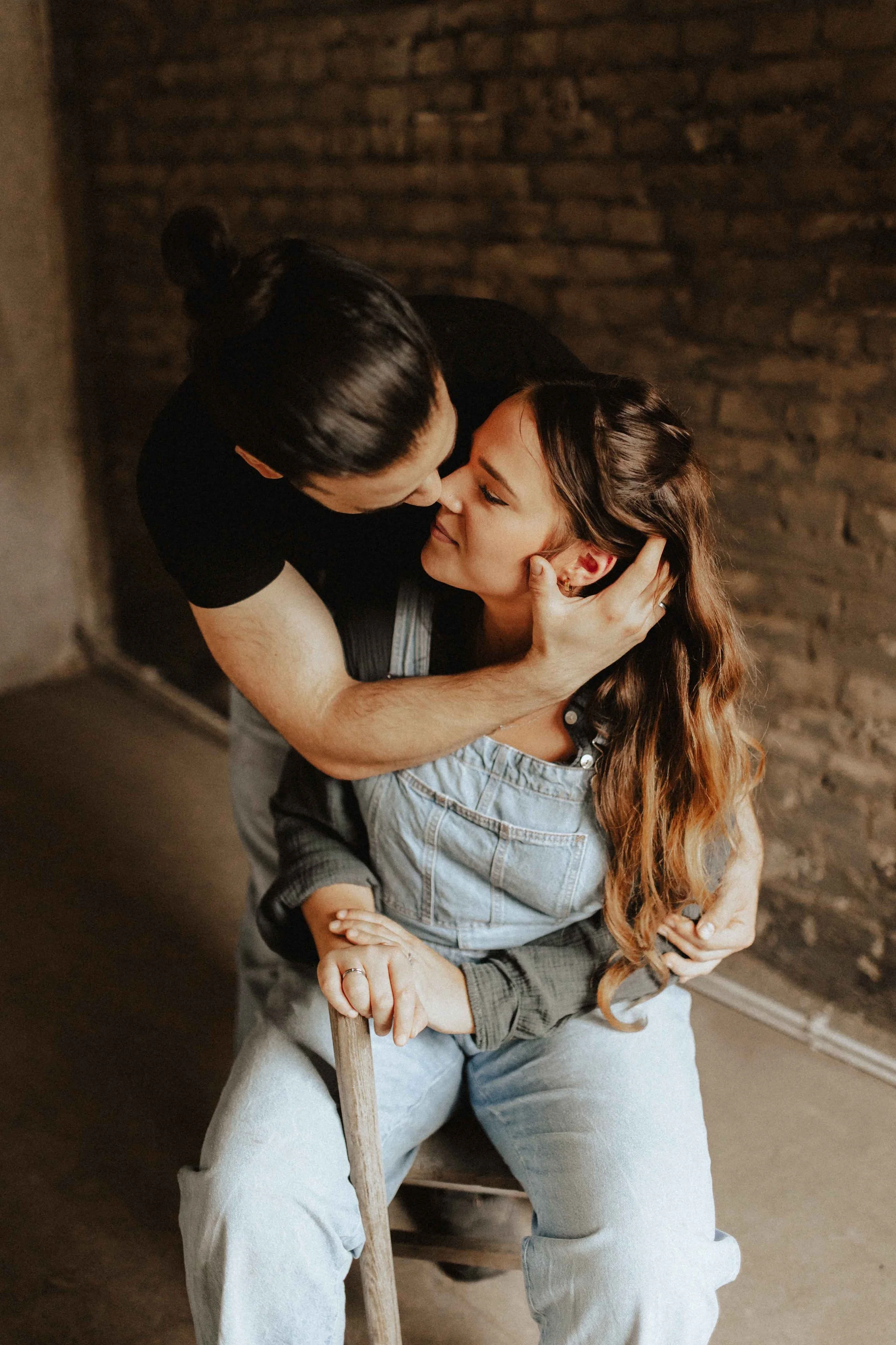 Zärtlicher Moment bei einem Paarshooting in Lüneburg – Mann umarmt und küsst seine Partnerin auf einem Stuhl vor rustikaler Backsteinwand, authentische und emotionale Paarfotografie.