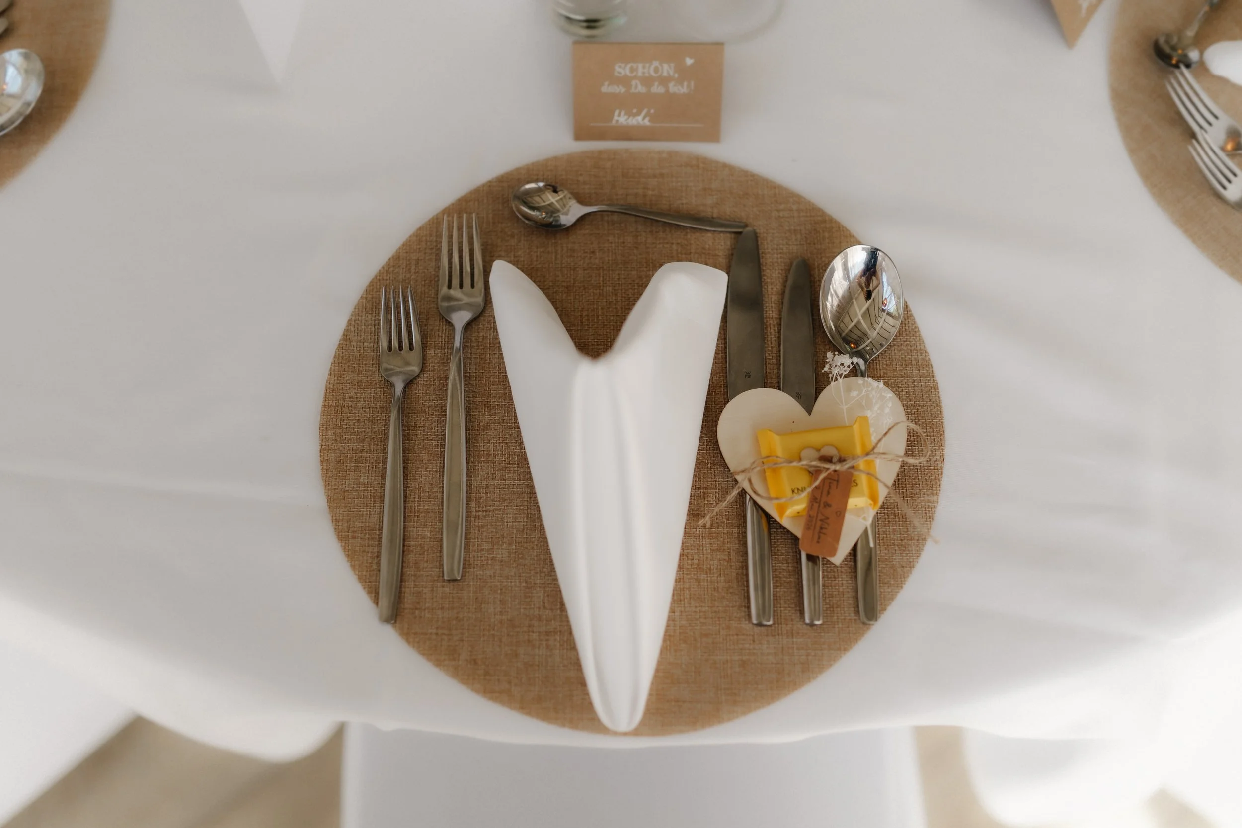 Detailaufnahme einer liebevoll gedeckten Hochzeitstafel mit herzförmig gefalteter weißer Serviette, rundem Jute-Platzset und kleinem Gastgeschenk in Herzform.