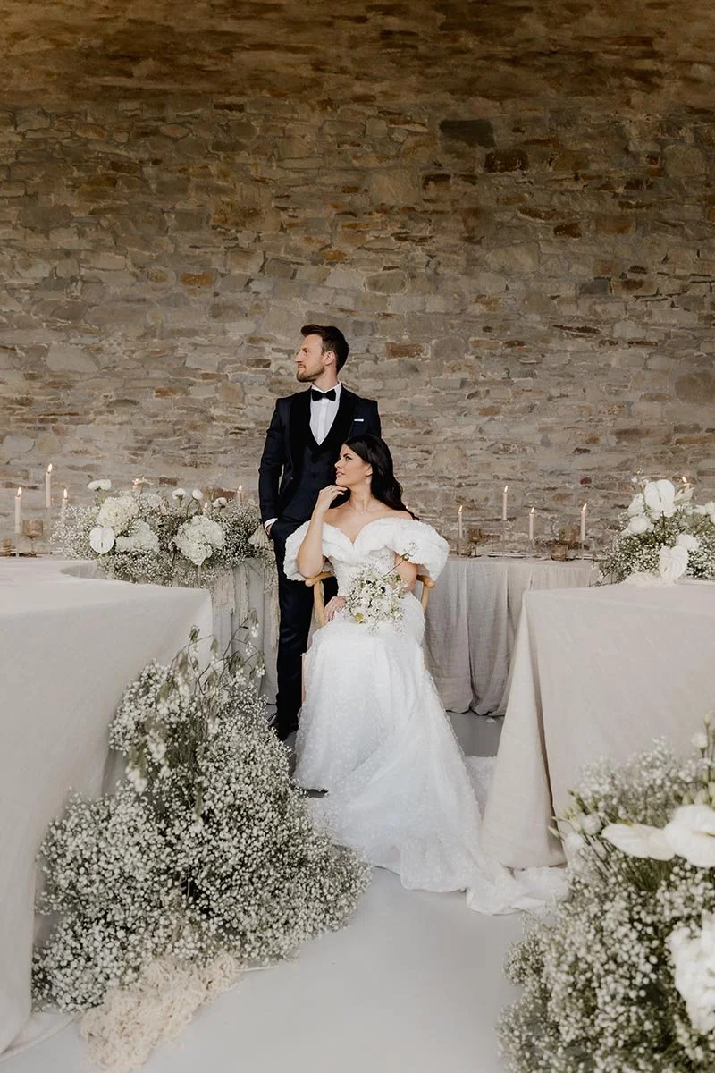 Brautpaar in eleganter Hochzeitskleidung vor einer rustikalen Steinwand, umgeben von weißen Blumen und einer festlich gedeckten Tafel