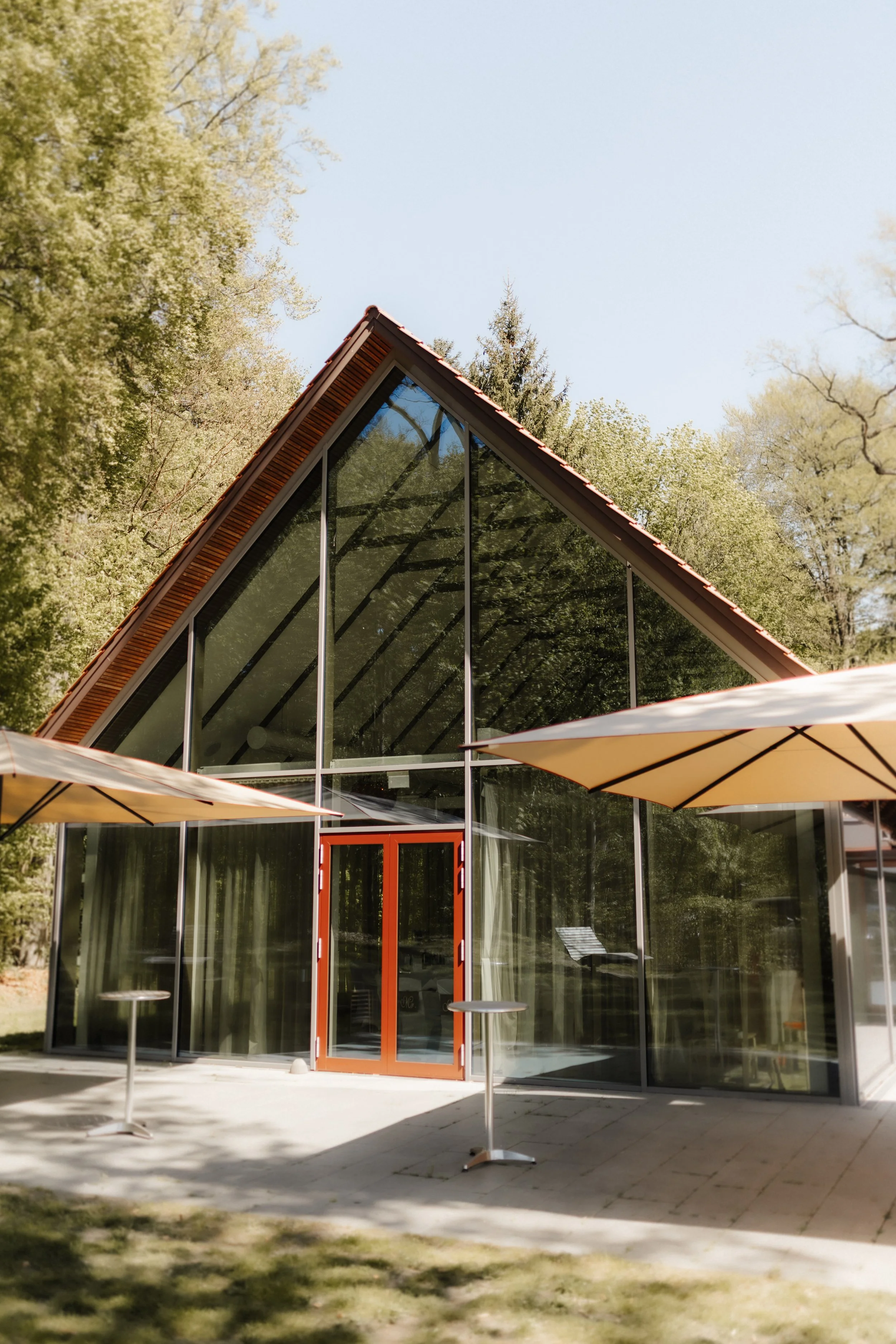 Moderne Hochzeitslocation mit großer Glasfront, spitzem Giebeldach und Terrasse mit Sonnenschirmen, umgeben von grünen Bäumen.
