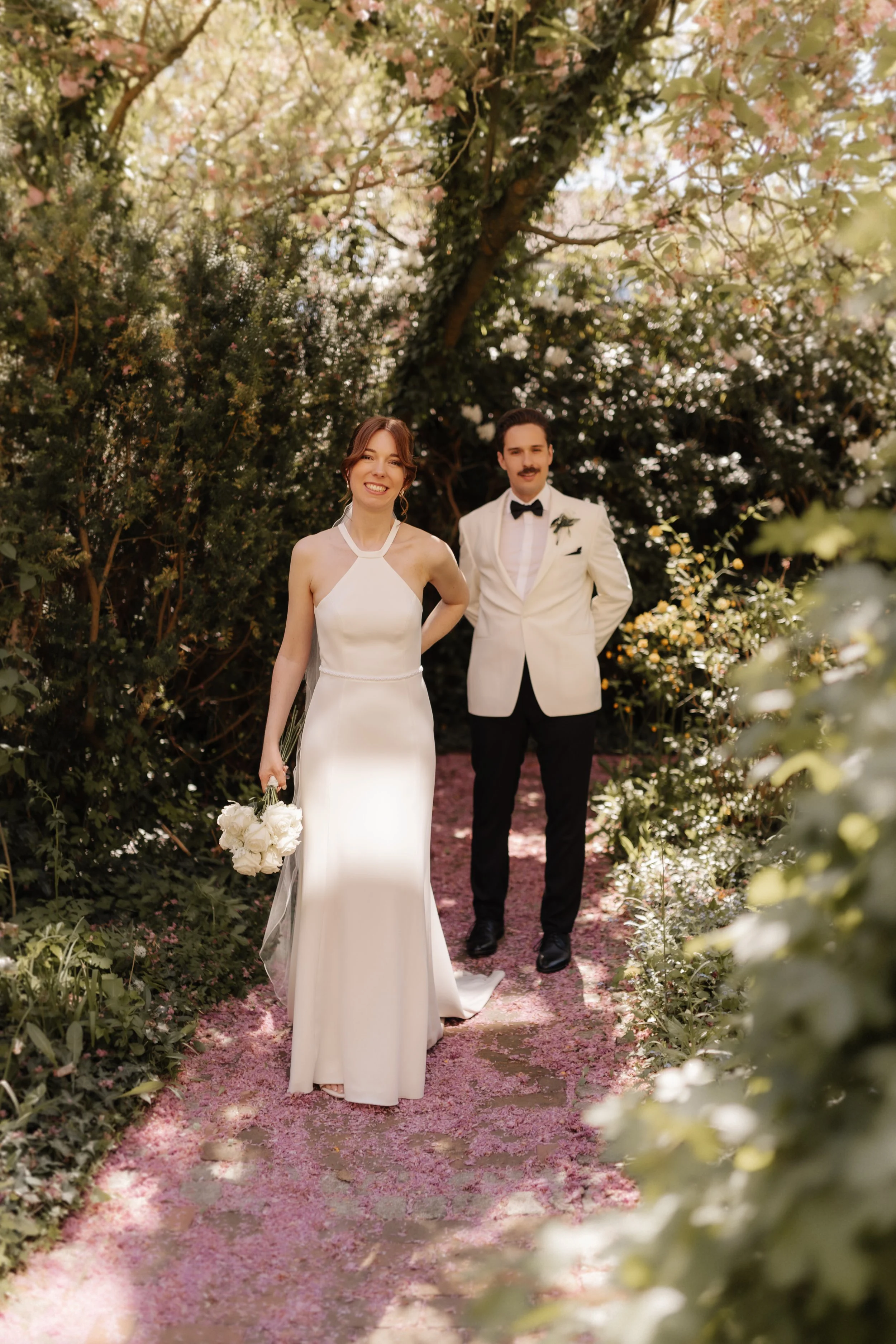 Braut und Bräutigam beim natürlichen Hochzeitsshooting im Garten des Brömsehauses in Lüneburg. Das Paar steht auf einem mit rosa Blüten bedeckten Gartenweg.