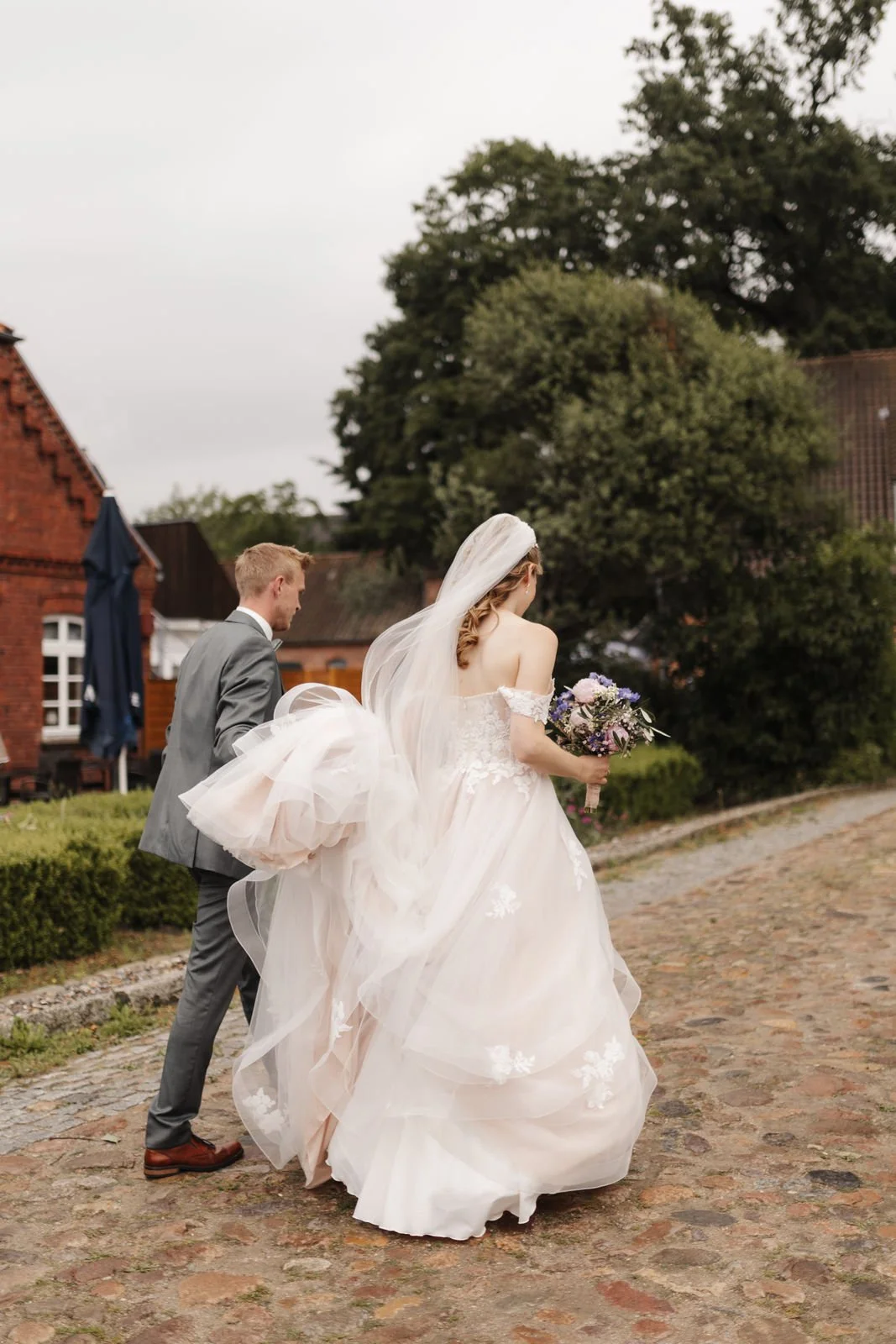 Brautpaar beim Spaziergang nach der Hochzeit im Heide Hotel Reinstorf bei Lüneburg. Der Bräutigam trägt die Schleppe des Brautkleides, während die Braut ihren Brautstrauß hält – natürliche Hochzeitsreportage.