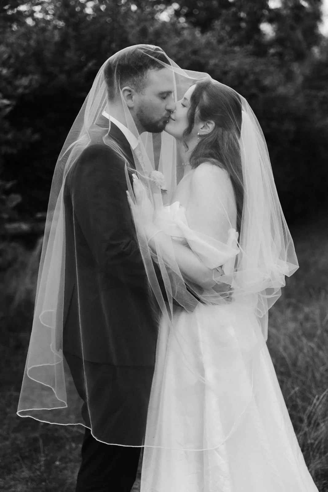Brautpaar küsst sich unter dem Brautschleier bei einem romantischen Paarshooting im Grünen – Hochzeitsfotografie in Schwarz-Weiß