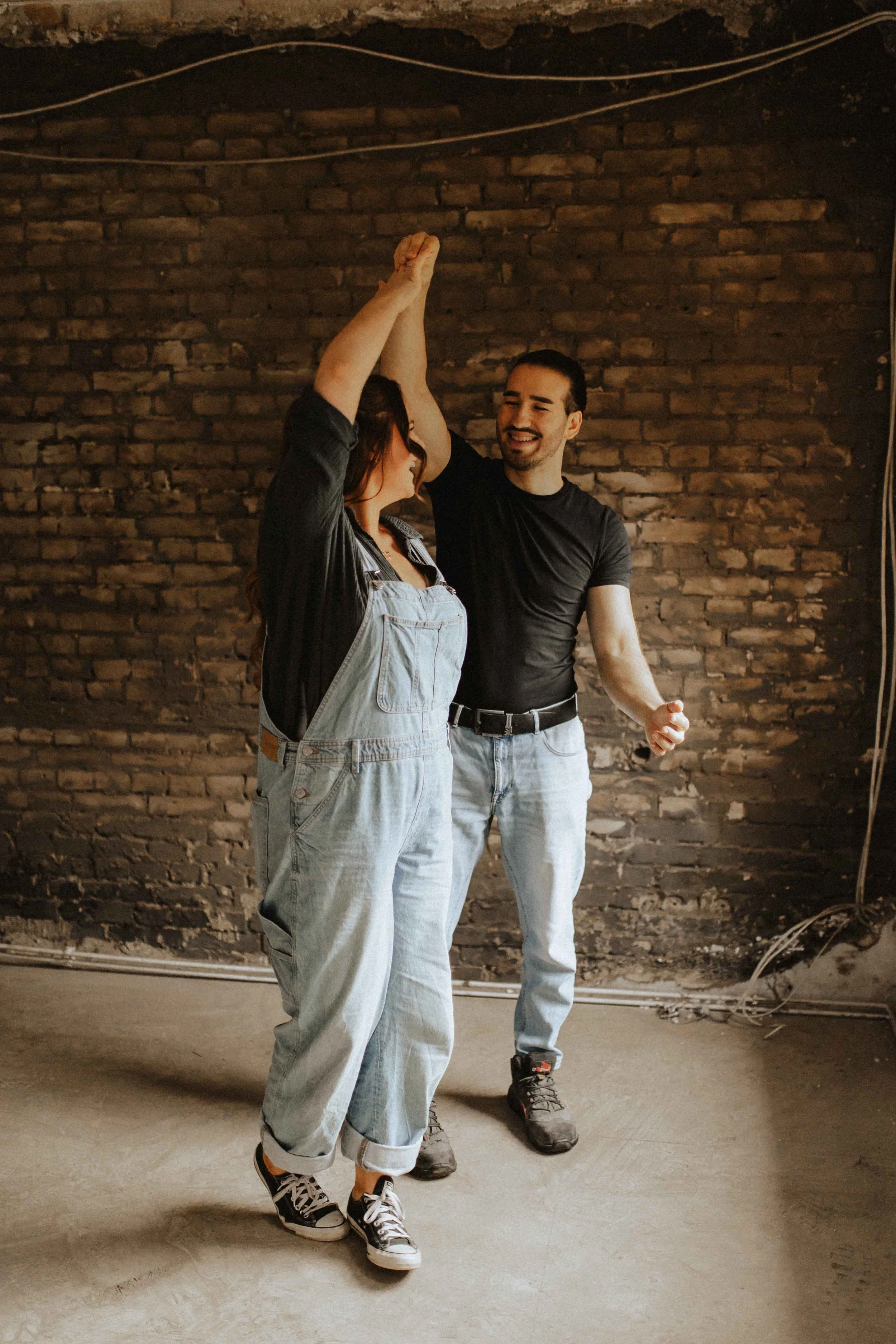 Baustellenshooting in Lüneburg – lachendes Paar tanzt gemeinsam vor einer Backsteinwand in Arbeitskleidung.  Du