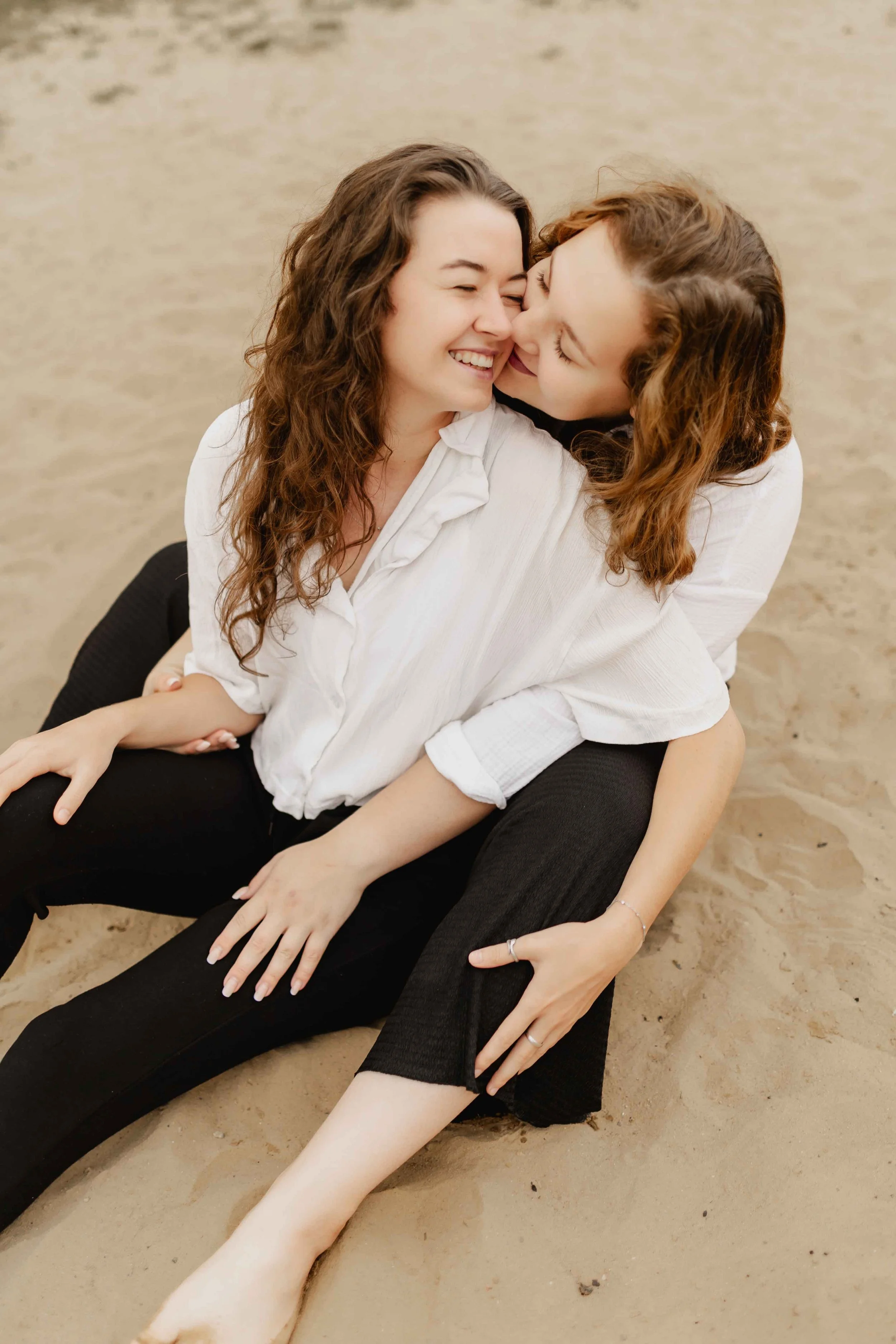 Zwei Frauen sitzen eng umschlungen im Sand, eine gibt der anderen einen liebevollen Kuss auf die Wange