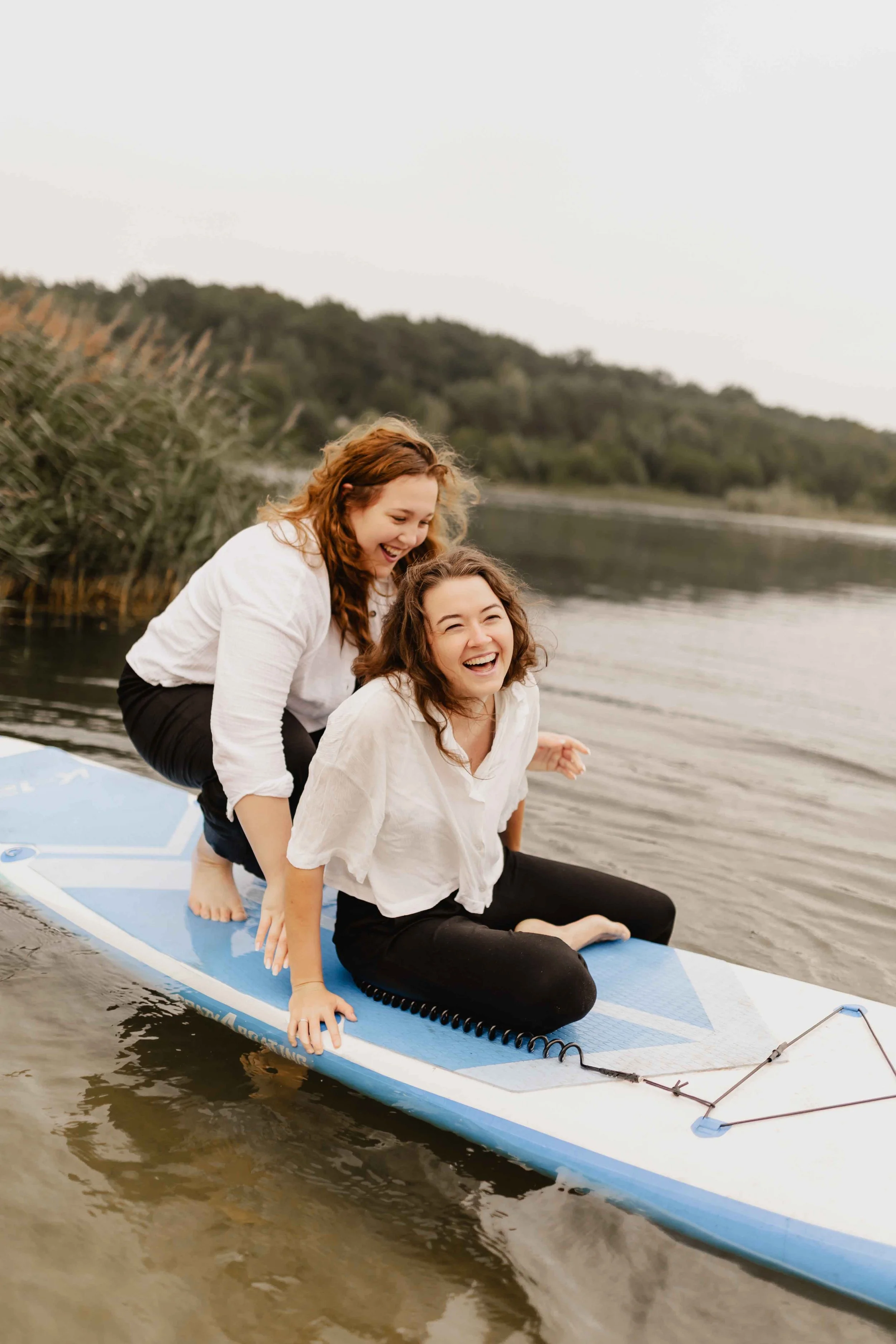 Zwei Frauen sitzen lachend zusammen auf einem SUP-Board und genießen den Moment am Wasser