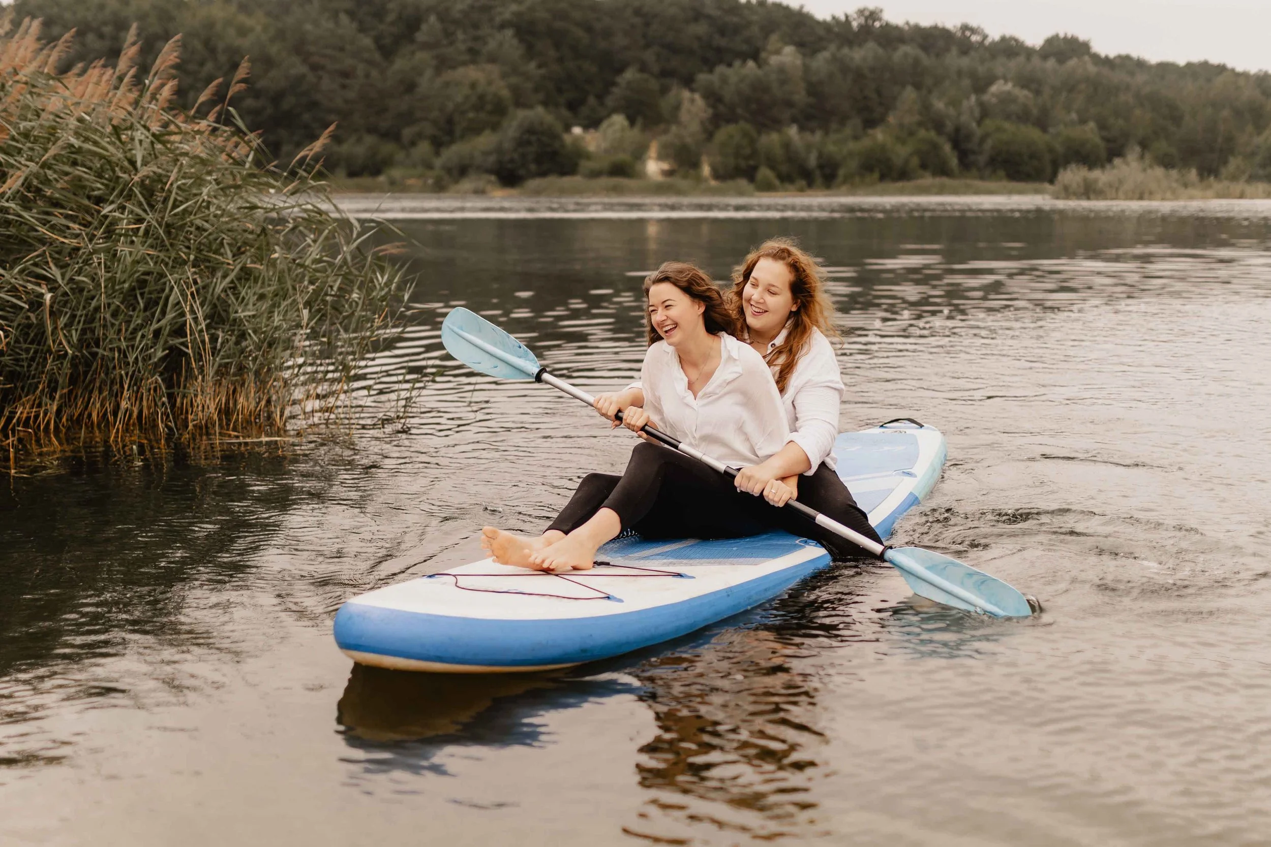 Zwei Frauen sitzen zusammen lachend auf einem SUP-Board und paddeln über den See