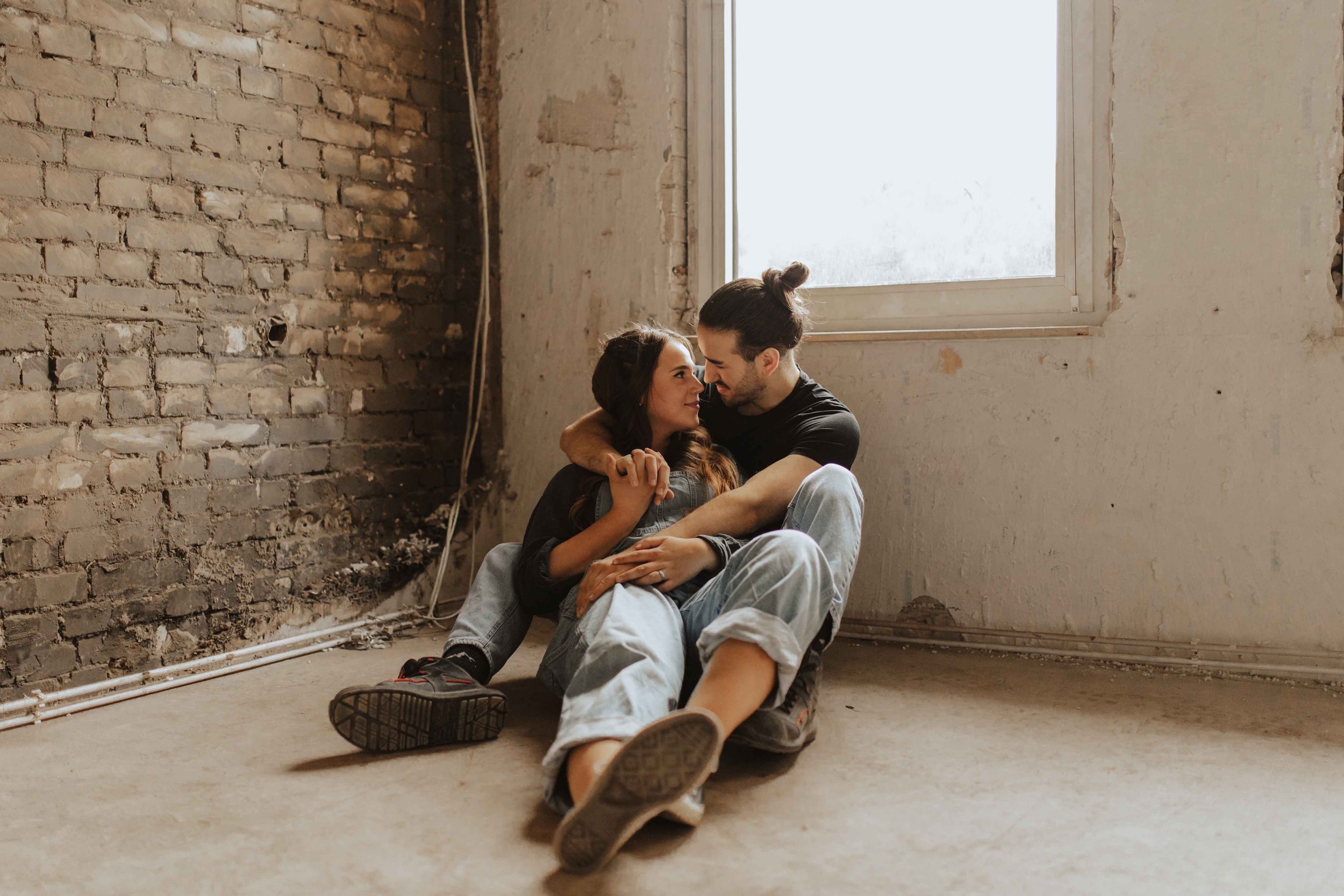 Authentisches Paarshooting in Lüneburg – verliebtes Paar sitzt eng umschlungen am Boden vor einer alten Backsteinwand, lächelnd und in inniger Umarmung.
