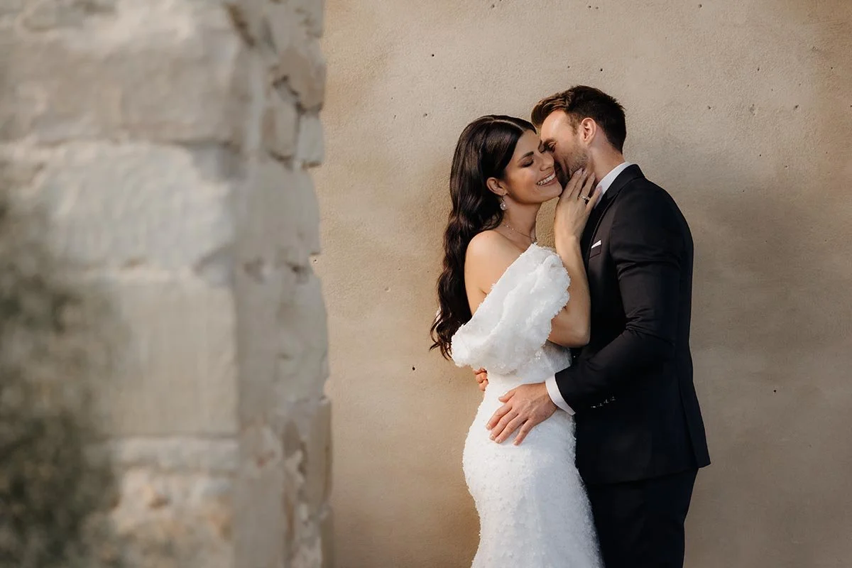 Brautpaar in eleganter Hochzeitskleidung bei einer innigen Umarmung vor einer hellen, rustikalen Wand