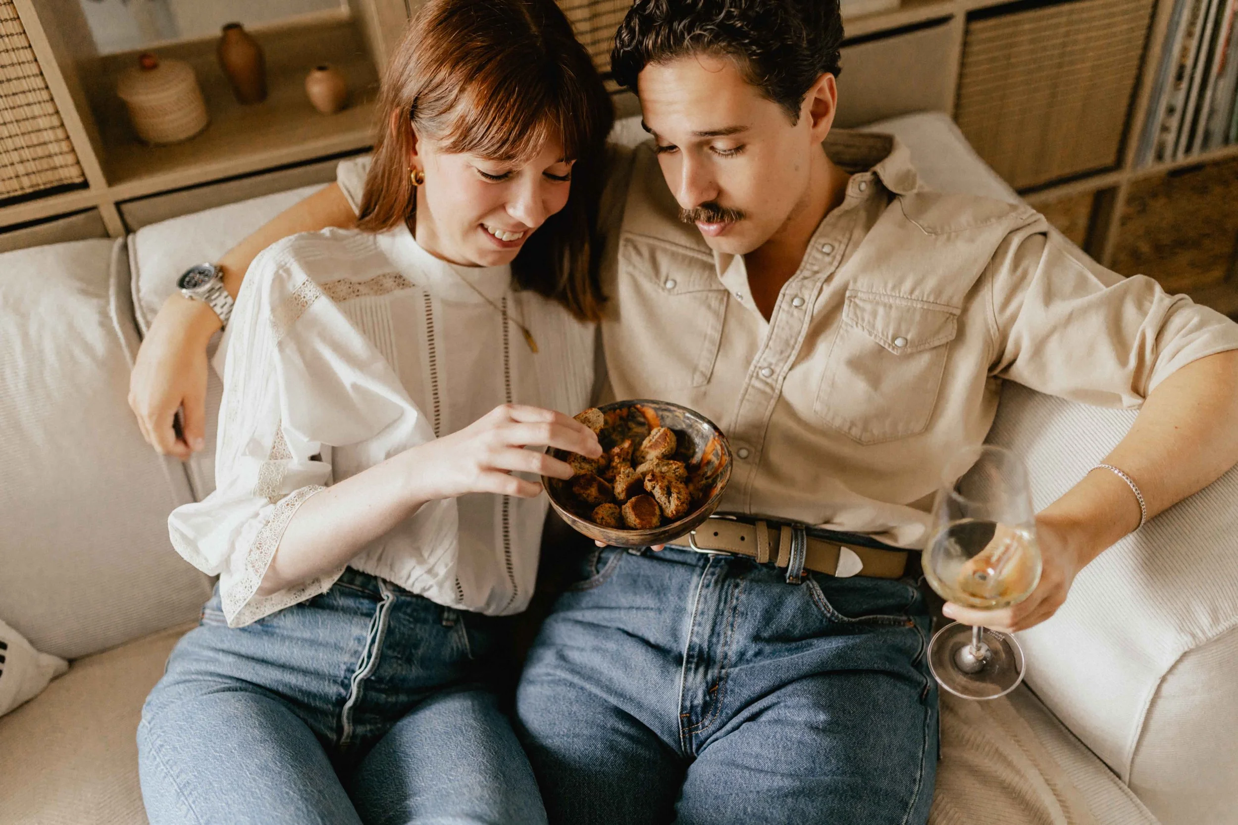 Paar sitzt bei einer Homestory in Lüneburg eng nebeneinander auf dem Sofa, teilt eine Schüssel mit Gebäck und trinkt Wein