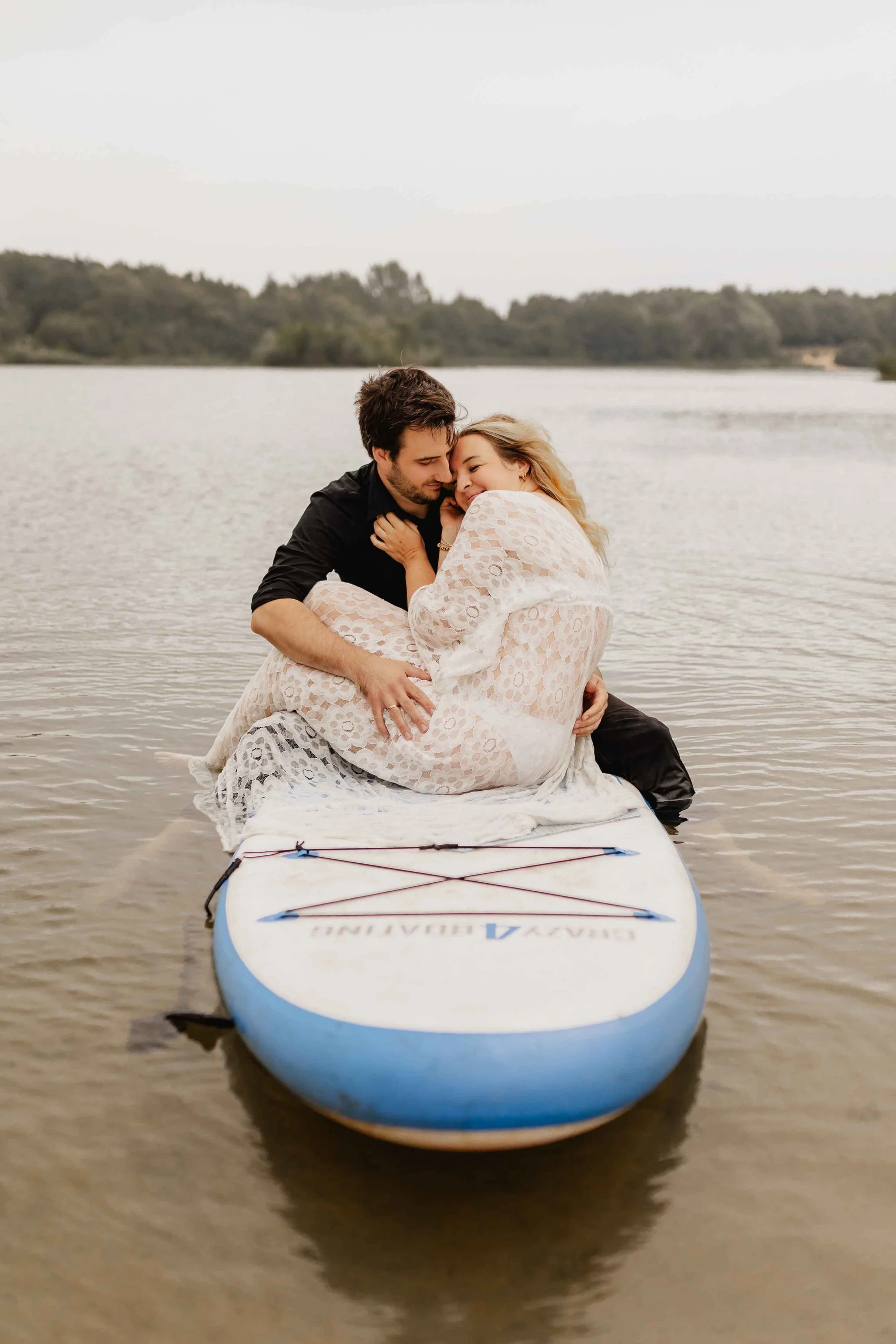 Paar sitzt eingekuschelt auf einem SUP-Board auf dem See und hält sich liebevoll im Arm