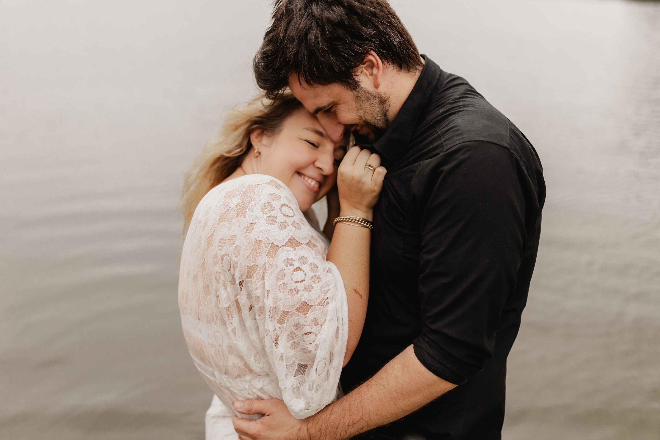 Paar steht eng umschlungen am See, sie lächelt vertrauensvoll an seiner Brust während beide Stirn an Stirn stehen