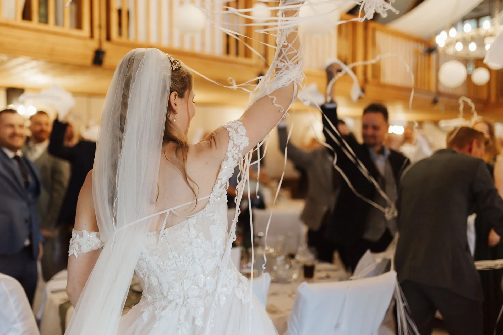 Farbfotografie einer Braut beim ausgelassenen Hochzeitsempfang im Heide Hotel Reinstorf bei Lüneburg, umgeben von feiernden Gästen und fliegenden Luftschlangen.