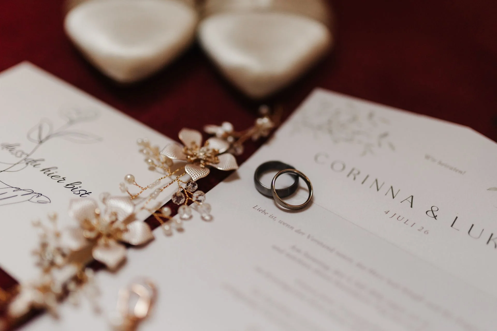 Romantische Detailaufnahme zweier Eheringe auf einer Hochzeitseinladung, umgeben von zartem goldenem Haarschmuck und weißen Brautschuhen.