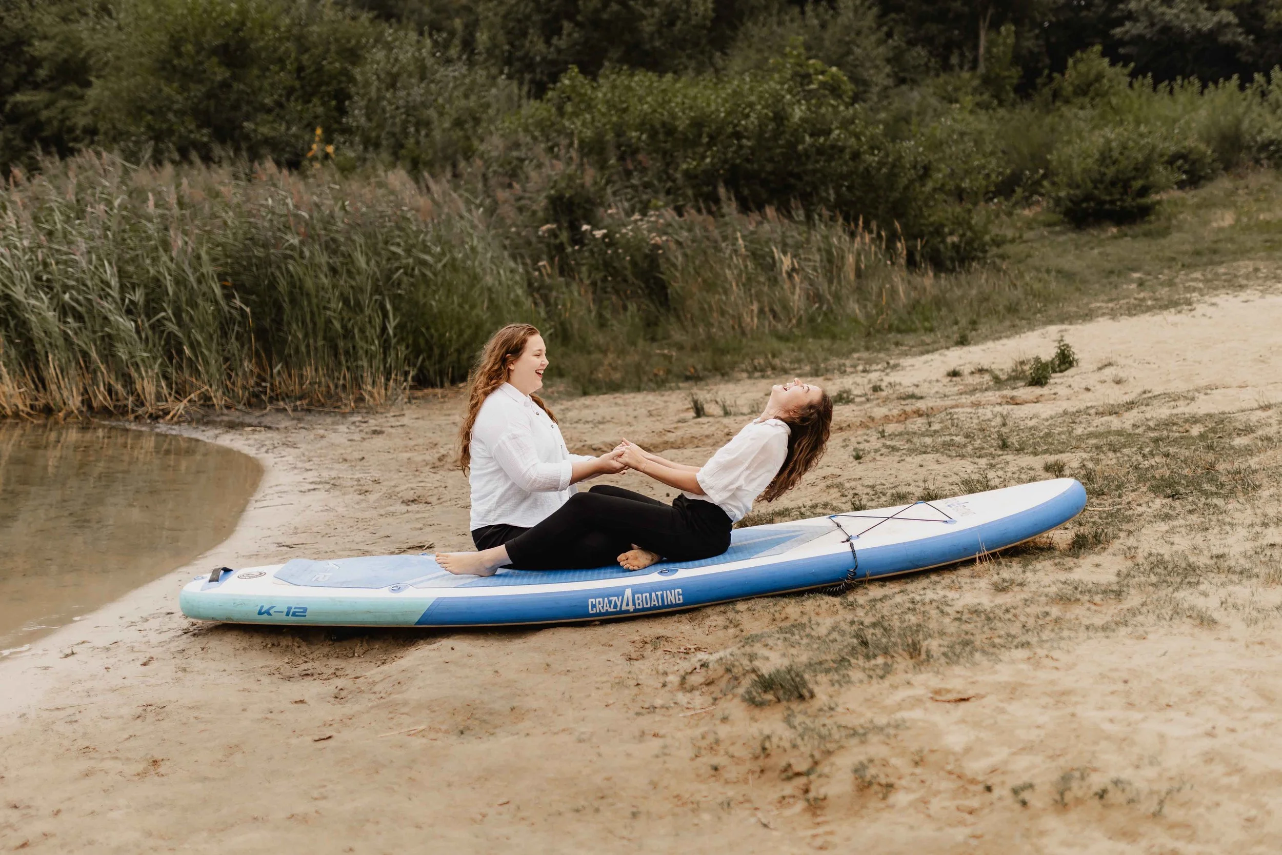 Zwei Frauen sitzen lachend auf einem SUP-Board am Ufer des Oldenstädter Sees