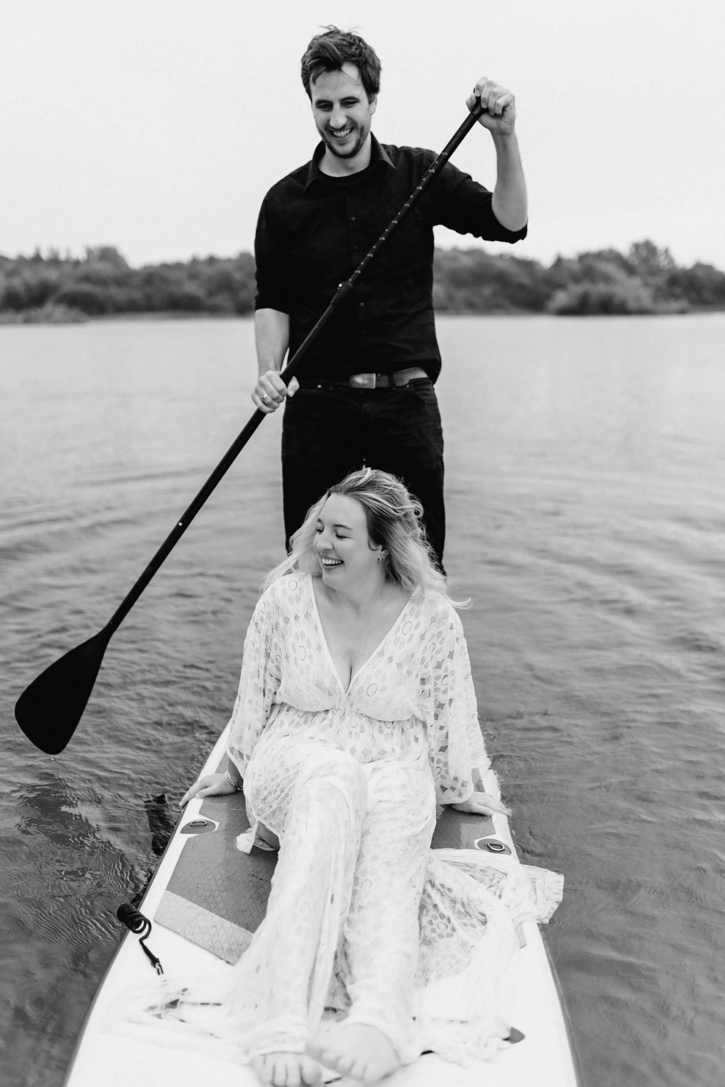 Schwarz-Weiß-Foto von einem Paar auf dem SUP-Board, er paddelt stehend während sie vorne lachend sitzt