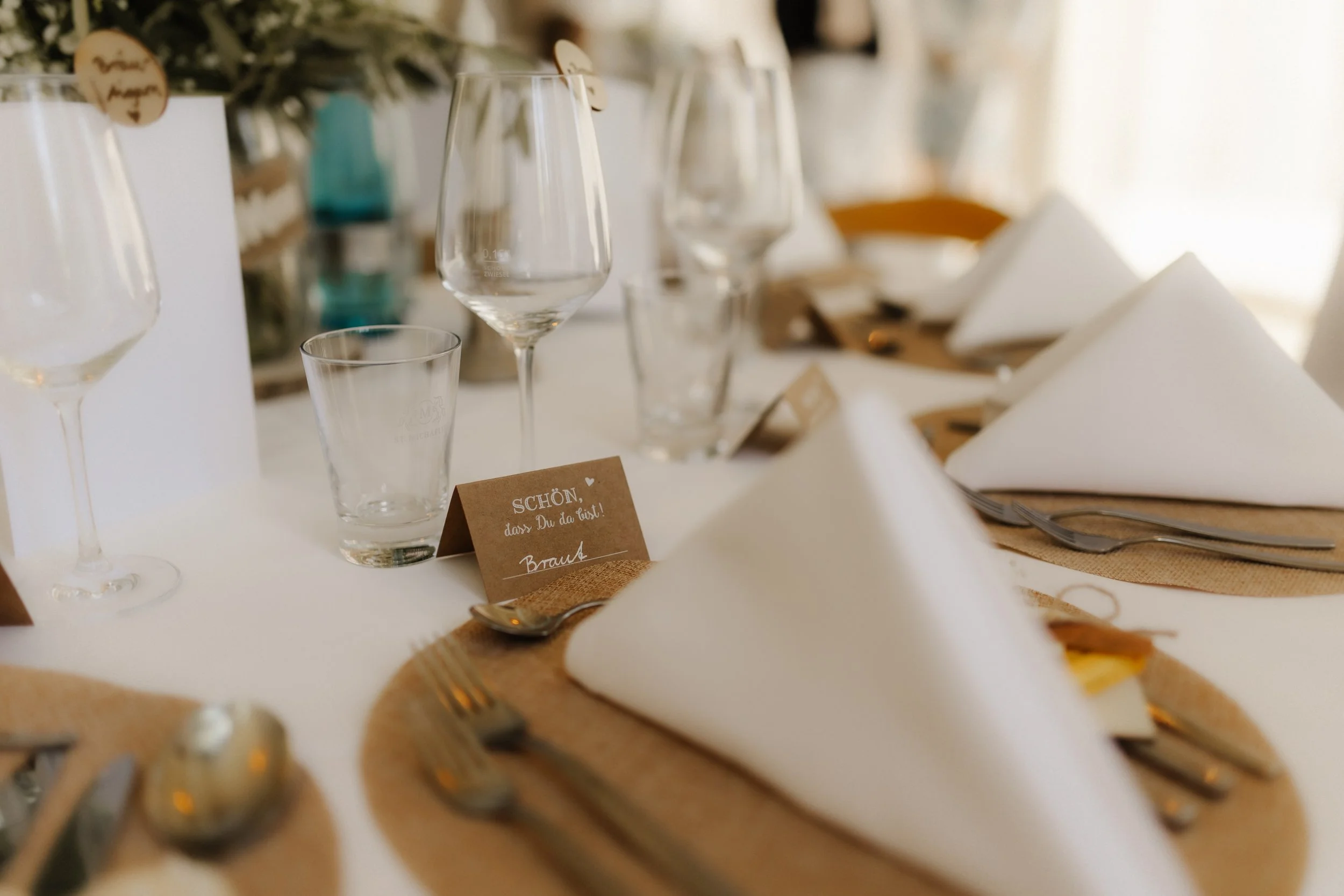Liebevoll gedeckte Hochzeitstafel mit weißen Servietten, Weingläsern, Jute-Platzsets und rustikalen Tischkarten bei einer Hochzeitsfeier.