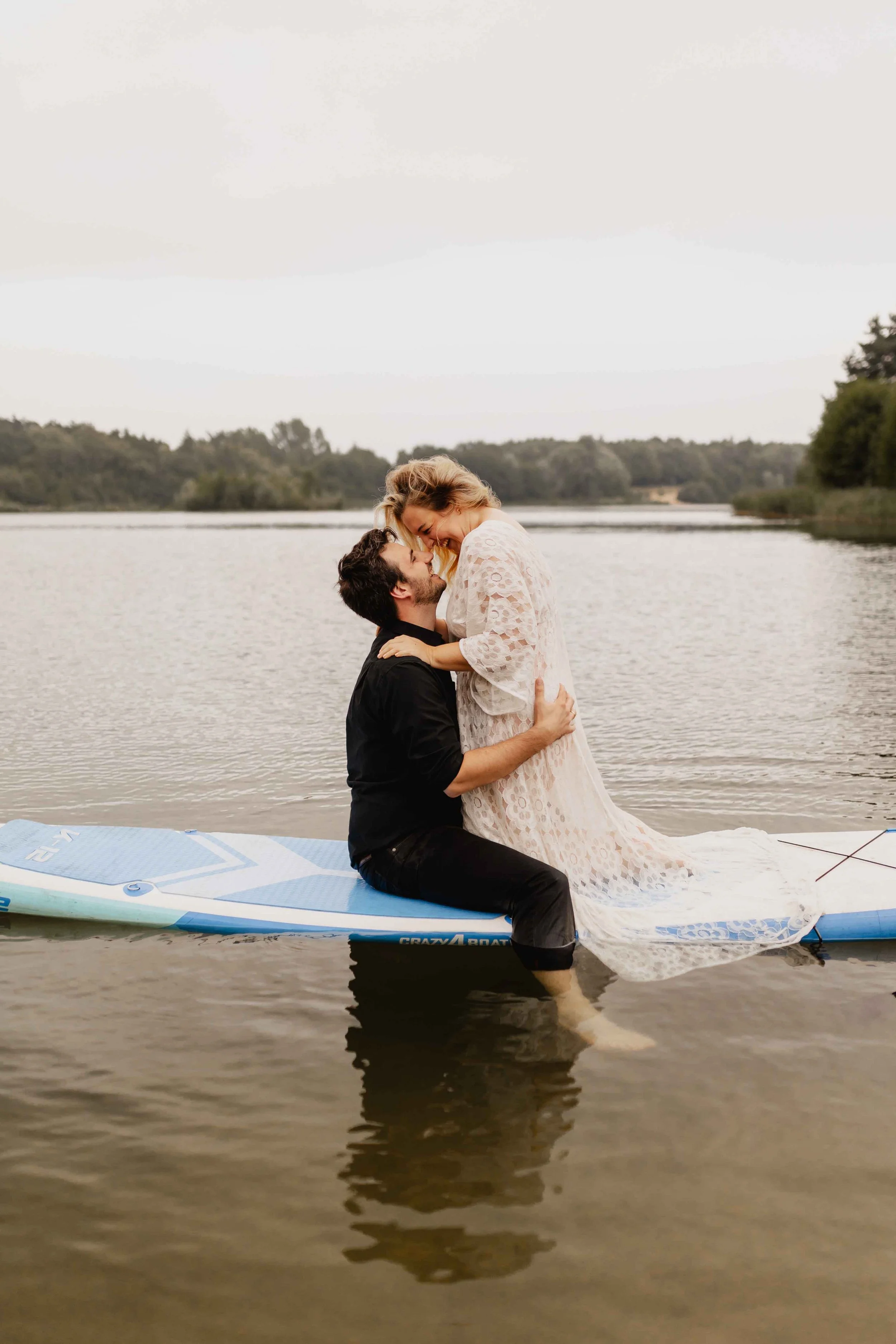 Paar sitzt auf SUP-Board am See, er hebt sie an und beide kuscheln sich in eine Decke