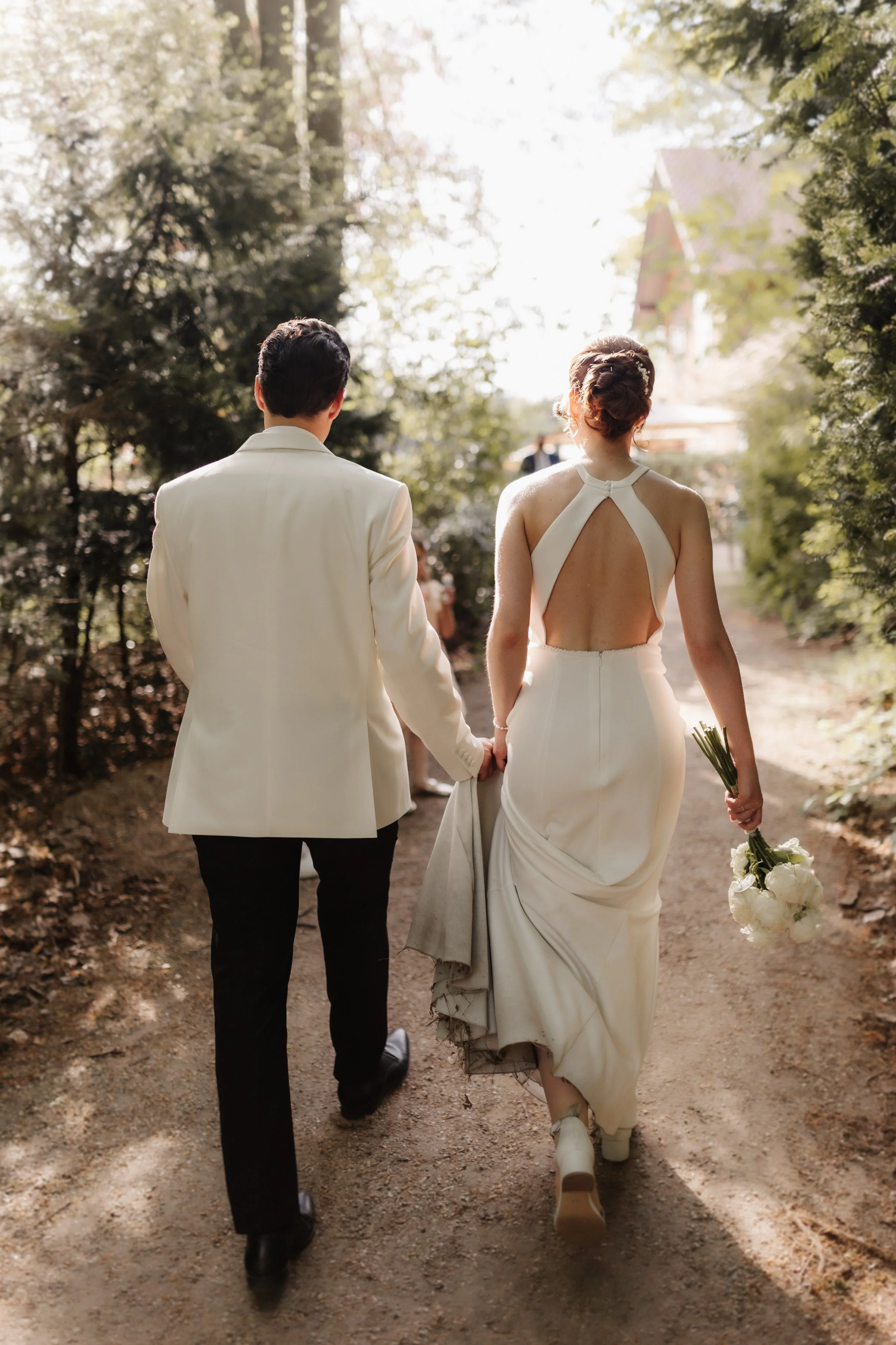 Brautpaar spaziert Hand in Hand über einen sonnigen Waldweg in Lüneburg. Die Braut trägt ein rückenfreies Hochzeitskleid und hält einen weißen Brautstrauß.